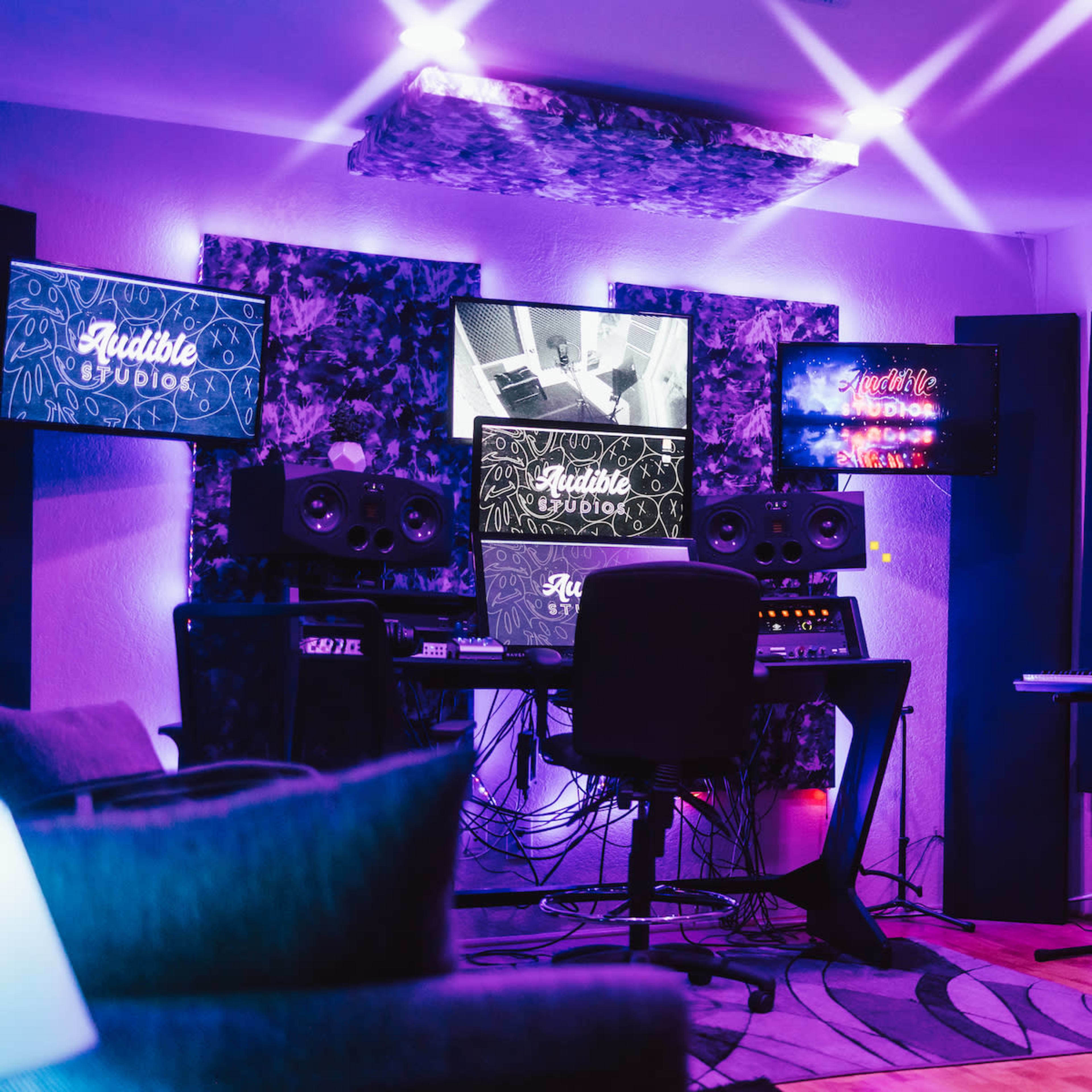 A music production studio features a central workstation with monitors, speakers, and equipment, surrounded by colorful lights and soundproofing materials.