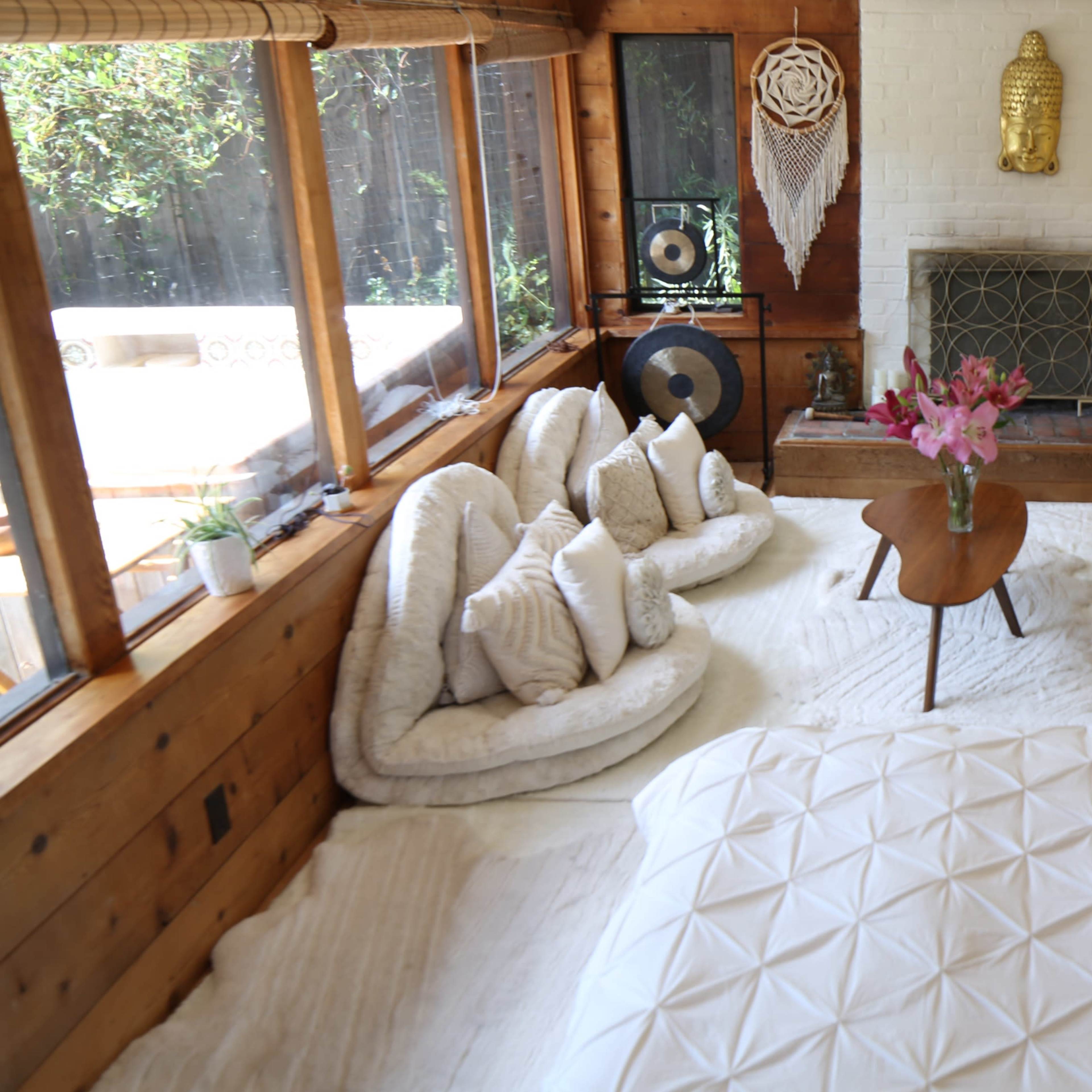 A cozy interior space featuring a white bed, a seating area with large cushions, a beige sofa, and decorative elements like plants and wall art.