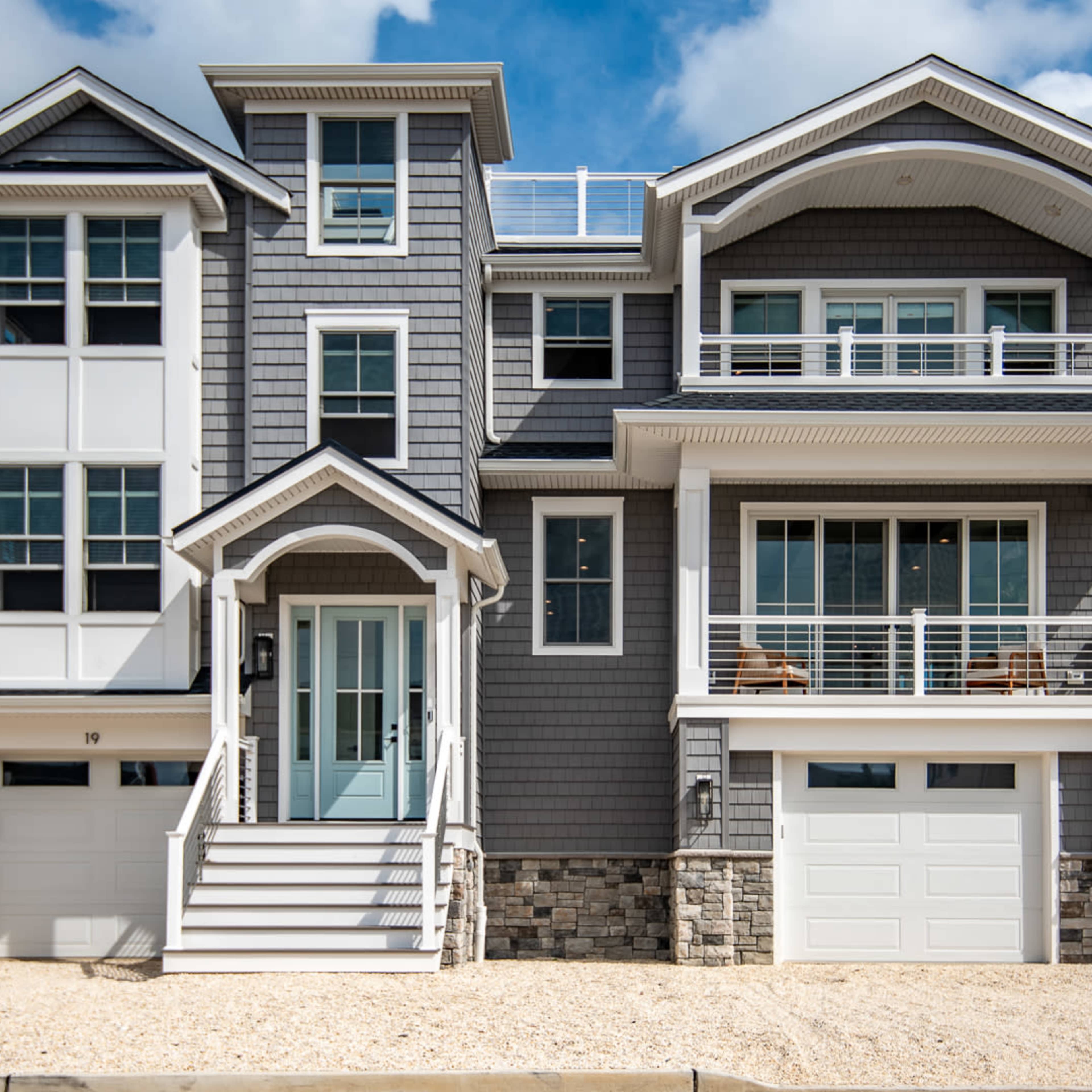 A modern gray-shingled house featuring multiple levels, large windows, and two garage doors, surrounded by a sandy area and a white fence.