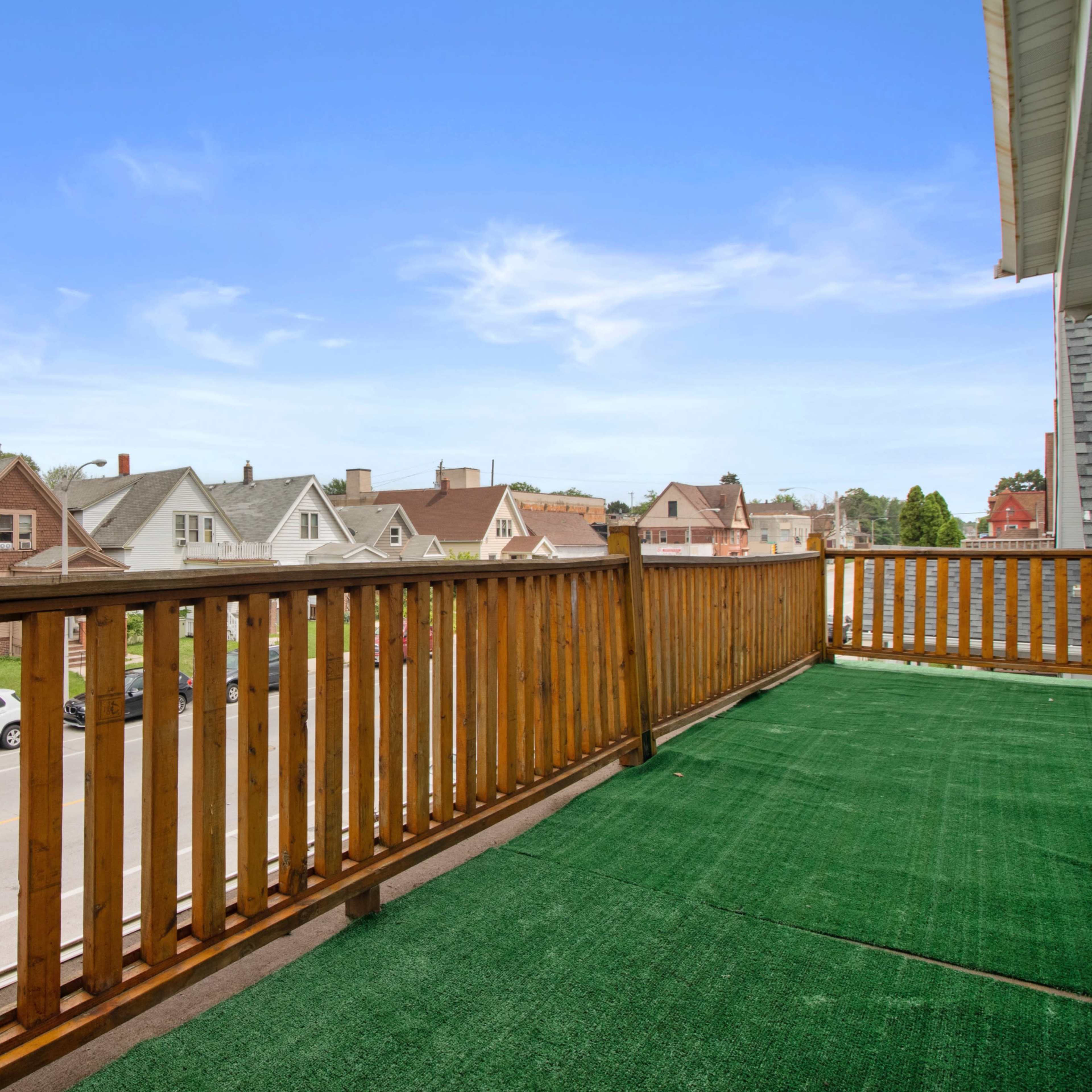 A wooden balcony with green carpeting overlooks a row of houses along a street.