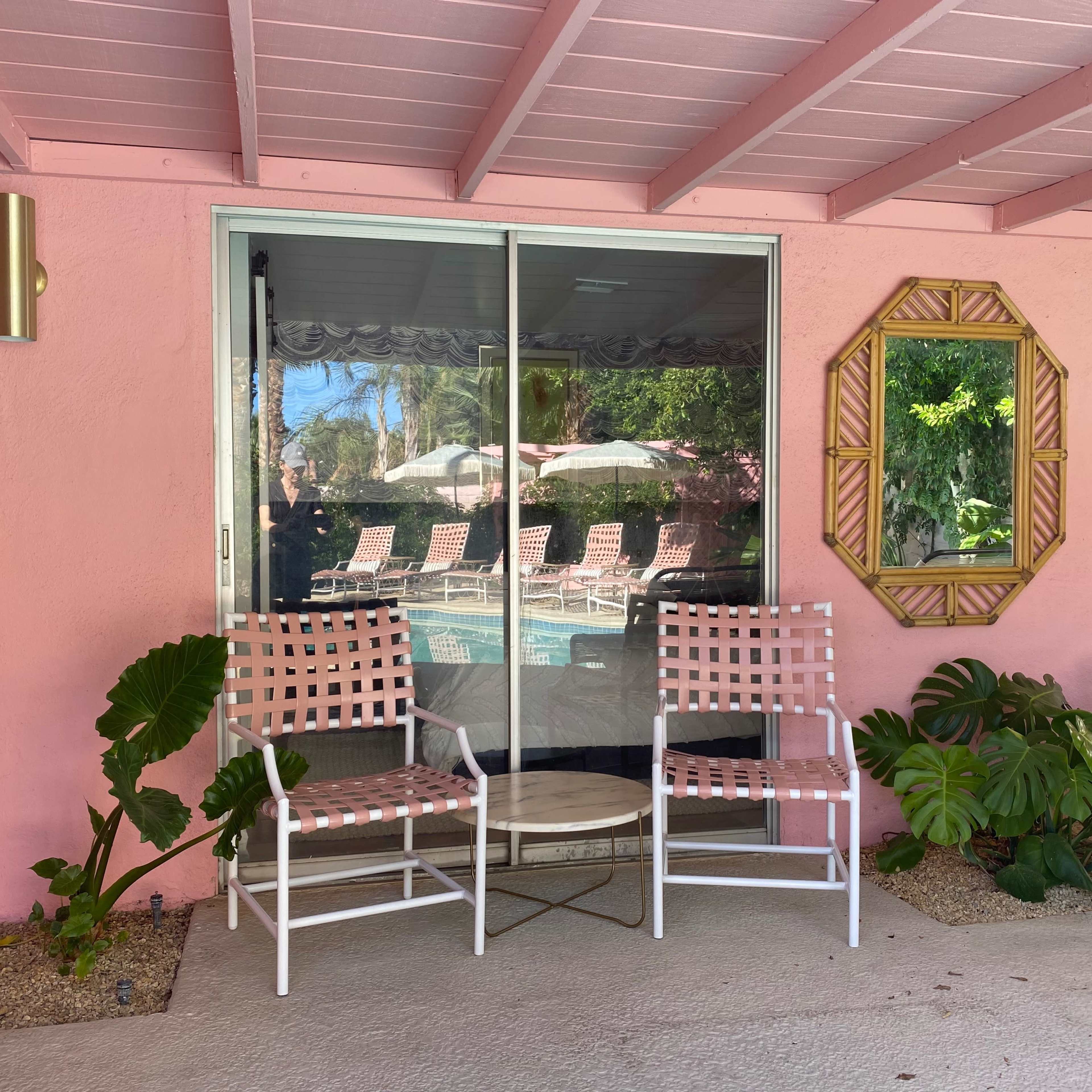 Two white chairs with pink accents and a small table are positioned outside a glass door that reflects a swimming pool area with loungers under umbrellas.