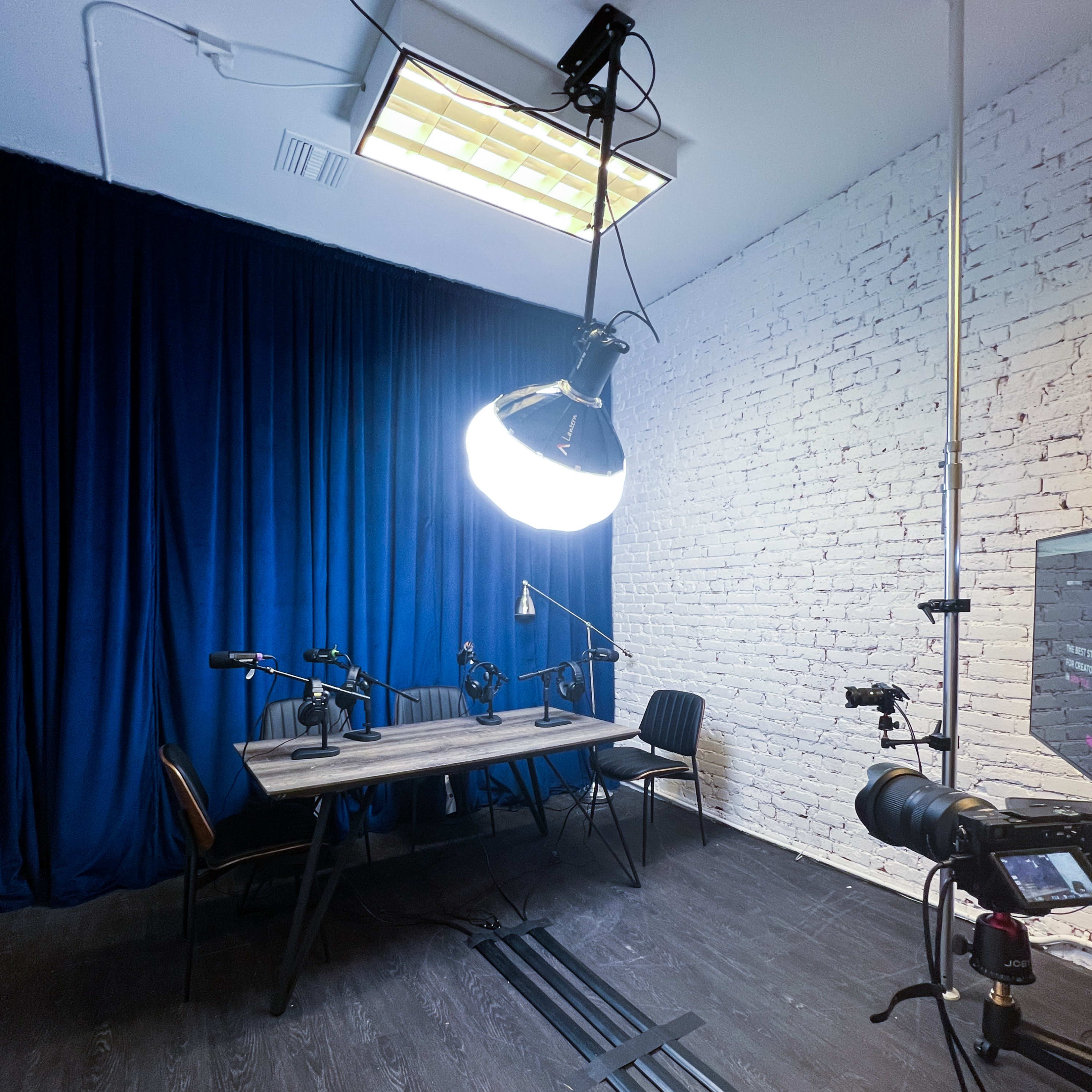A dimly lit recording studio features a large circular light above a table with multiple microphones and a chair, set against a backdrop of blue curtains and a white brick wall.