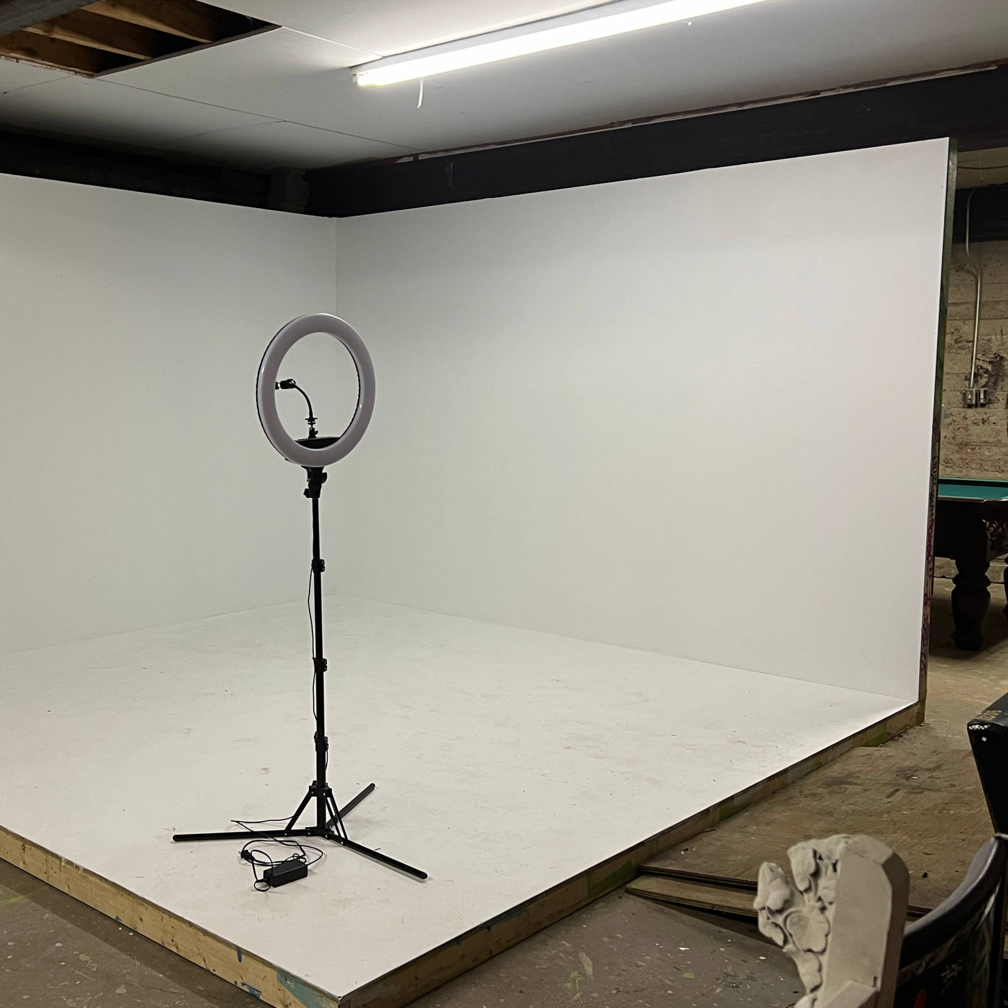 A plain, empty room with white walls and a ring light on a stand in the center.