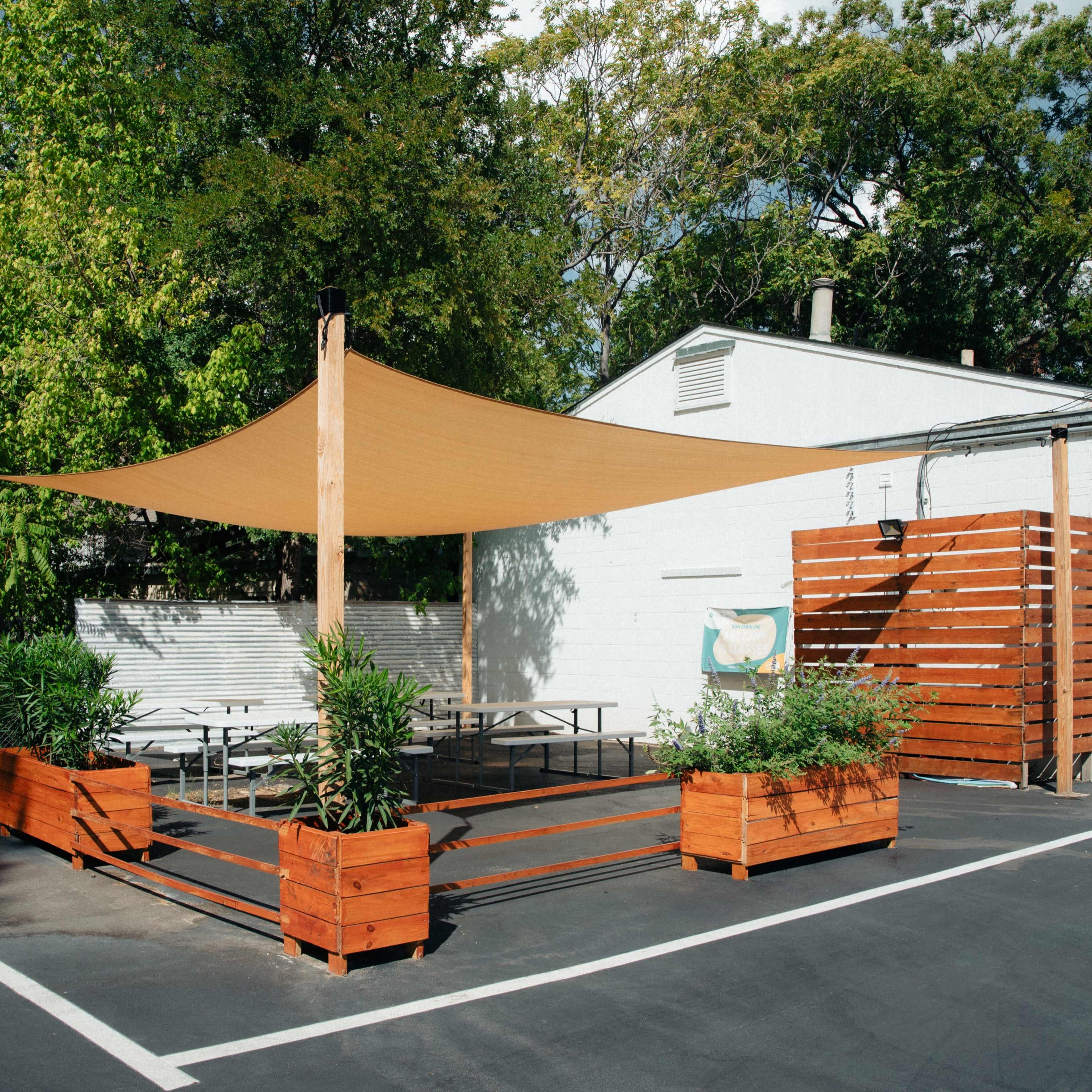 An outdoor seating area with wooden planters and a fabric shade structure, set against a backdrop of greenery and a white building.