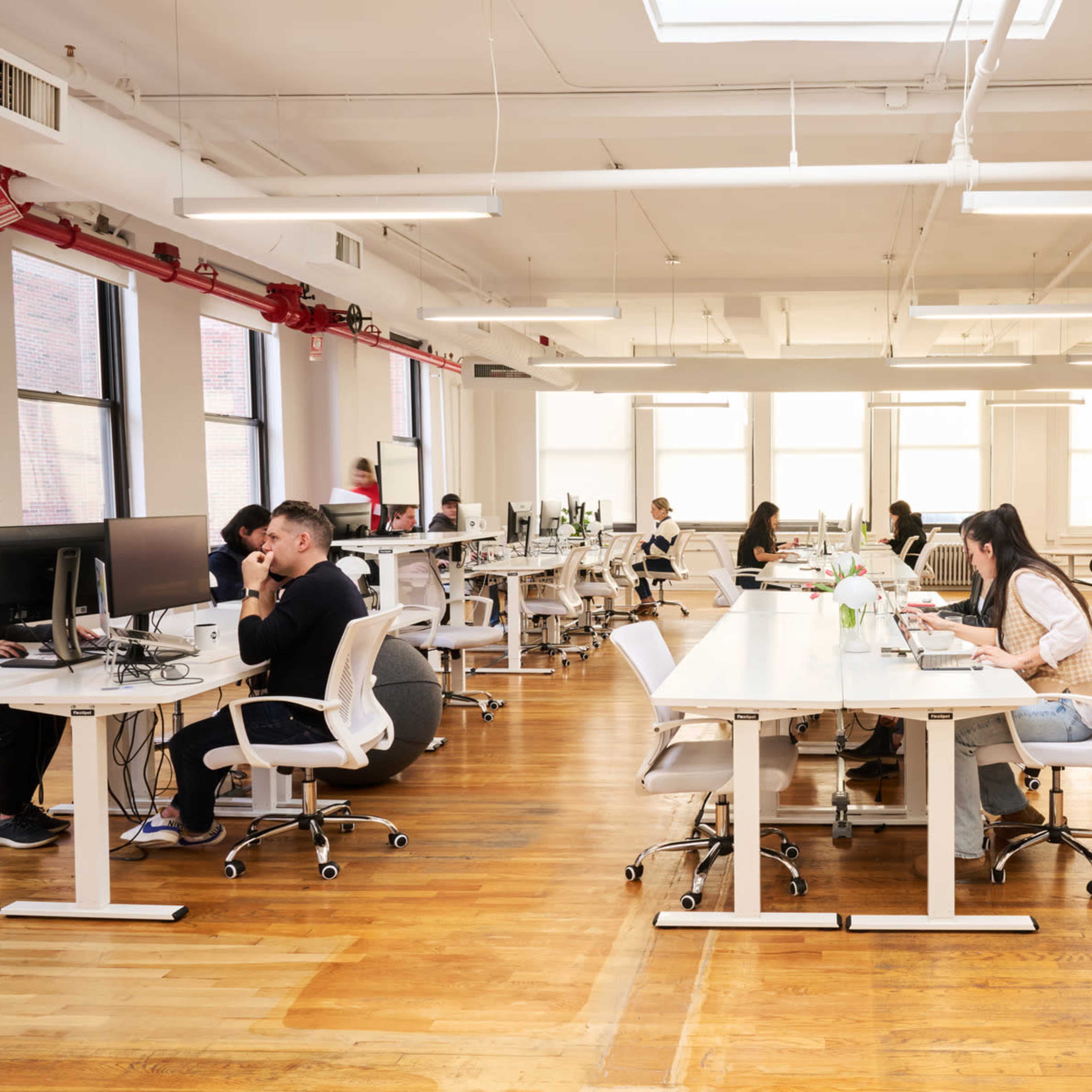 A modern office space features multiple workstations with people engaged in various tasks, surrounded by large windows and wooden flooring.