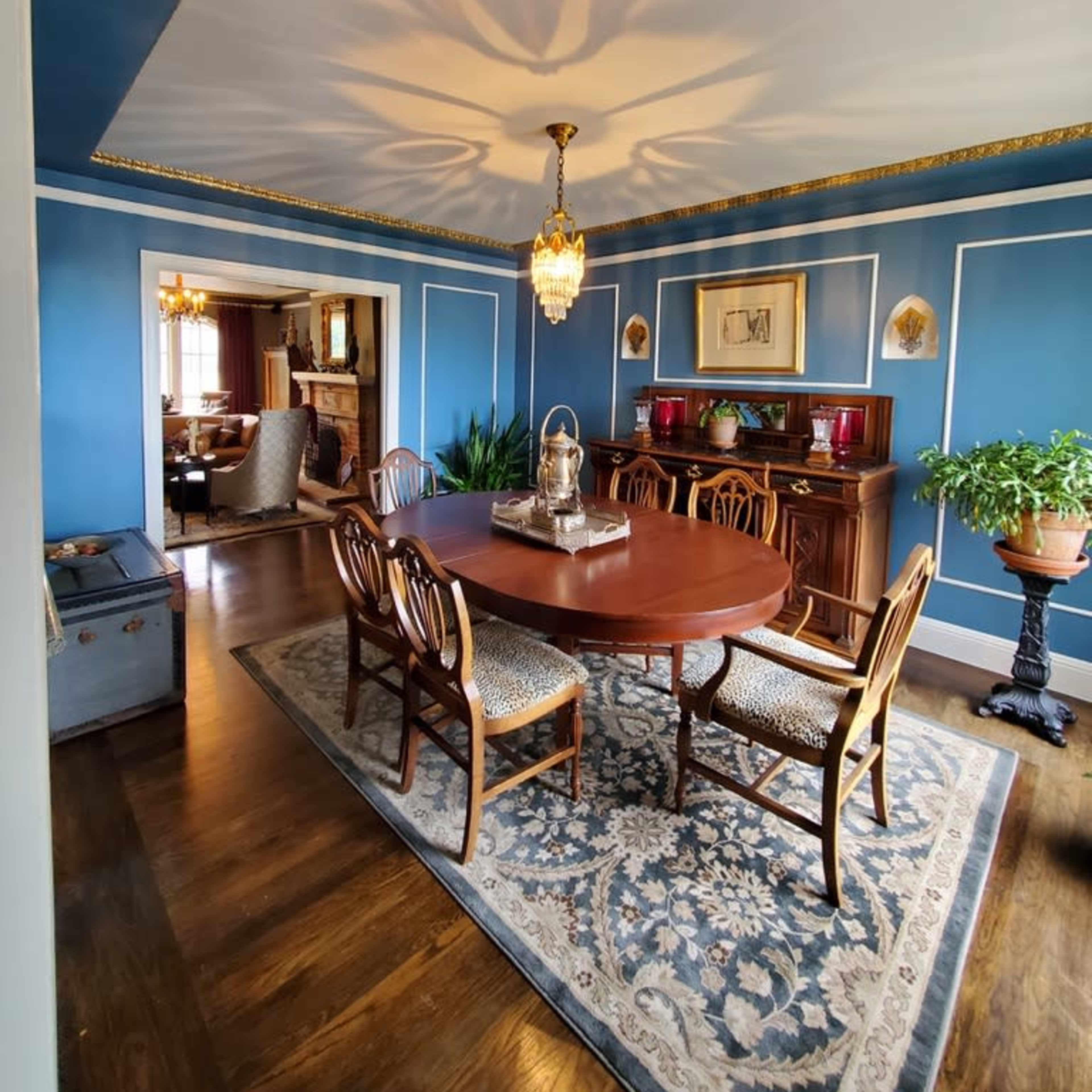 A dining room with a wooden table surrounded by chairs, blue walls, a chandelier, and a decorative rug.