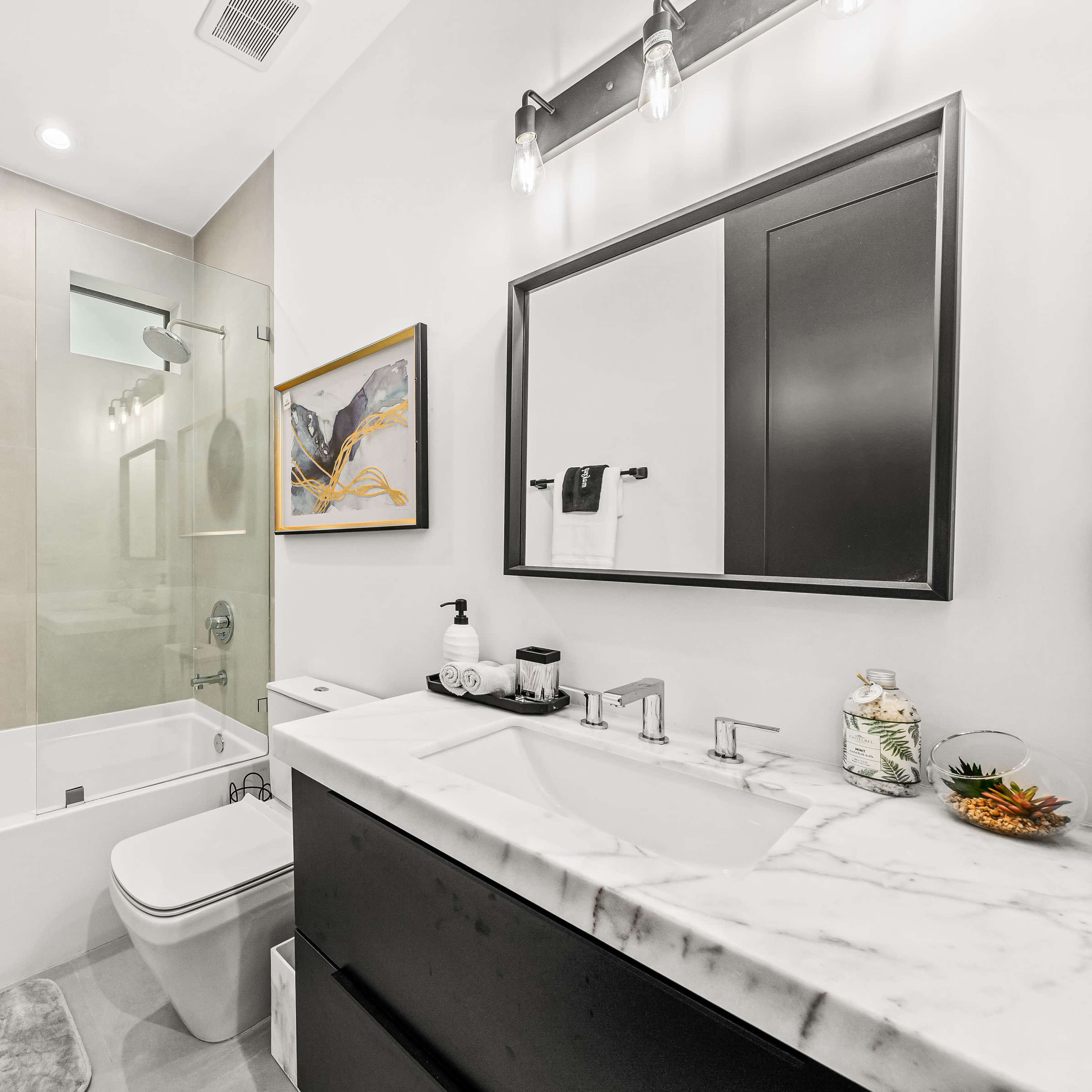 A modern bathroom featuring a bathtub and shower combination, a dual sink vanity with a marble countertop, and sleek black cabinetry.