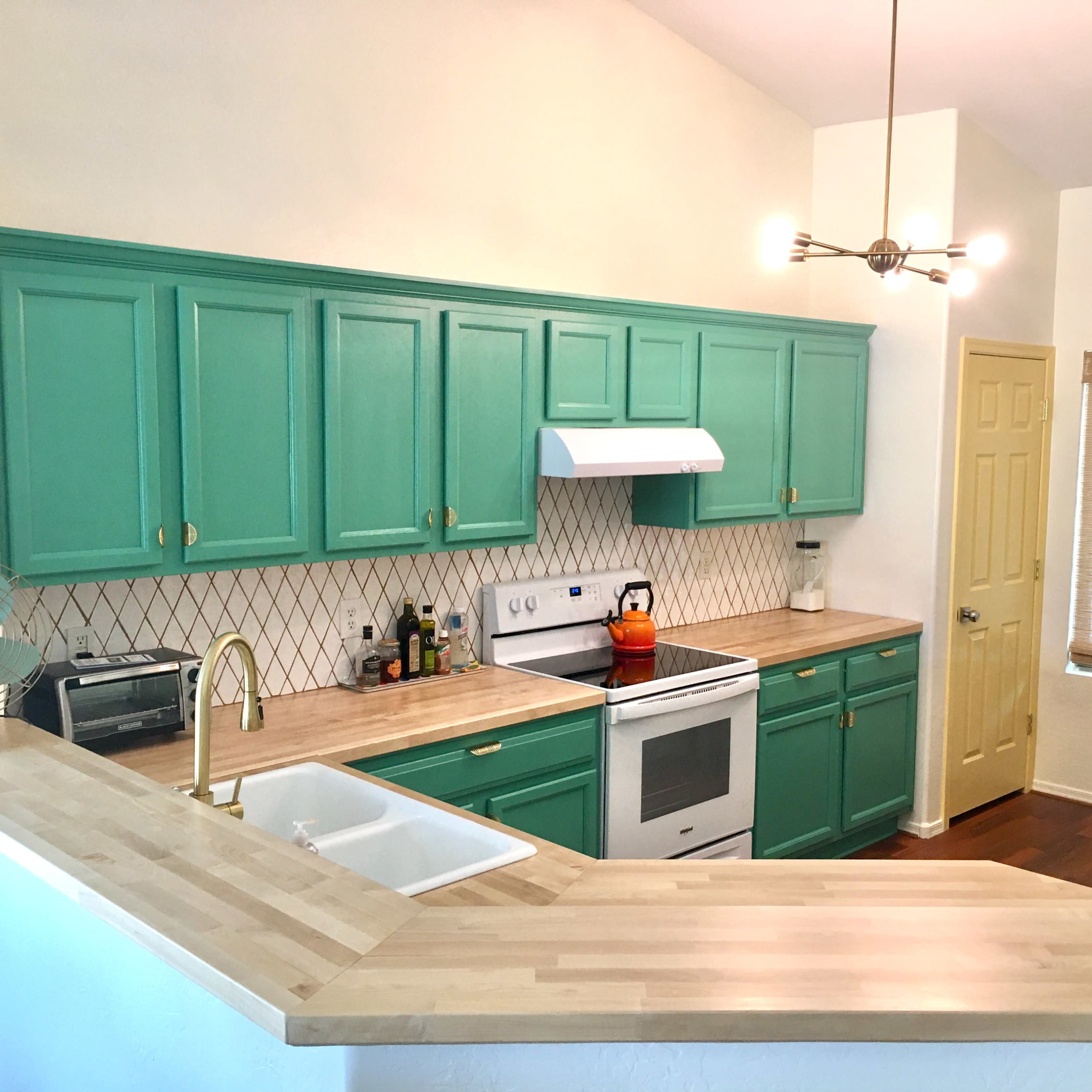 A modern kitchen with teal cabinets, a white stove and sink, and a stainless steel refrigerator, illuminated by overhead lighting.