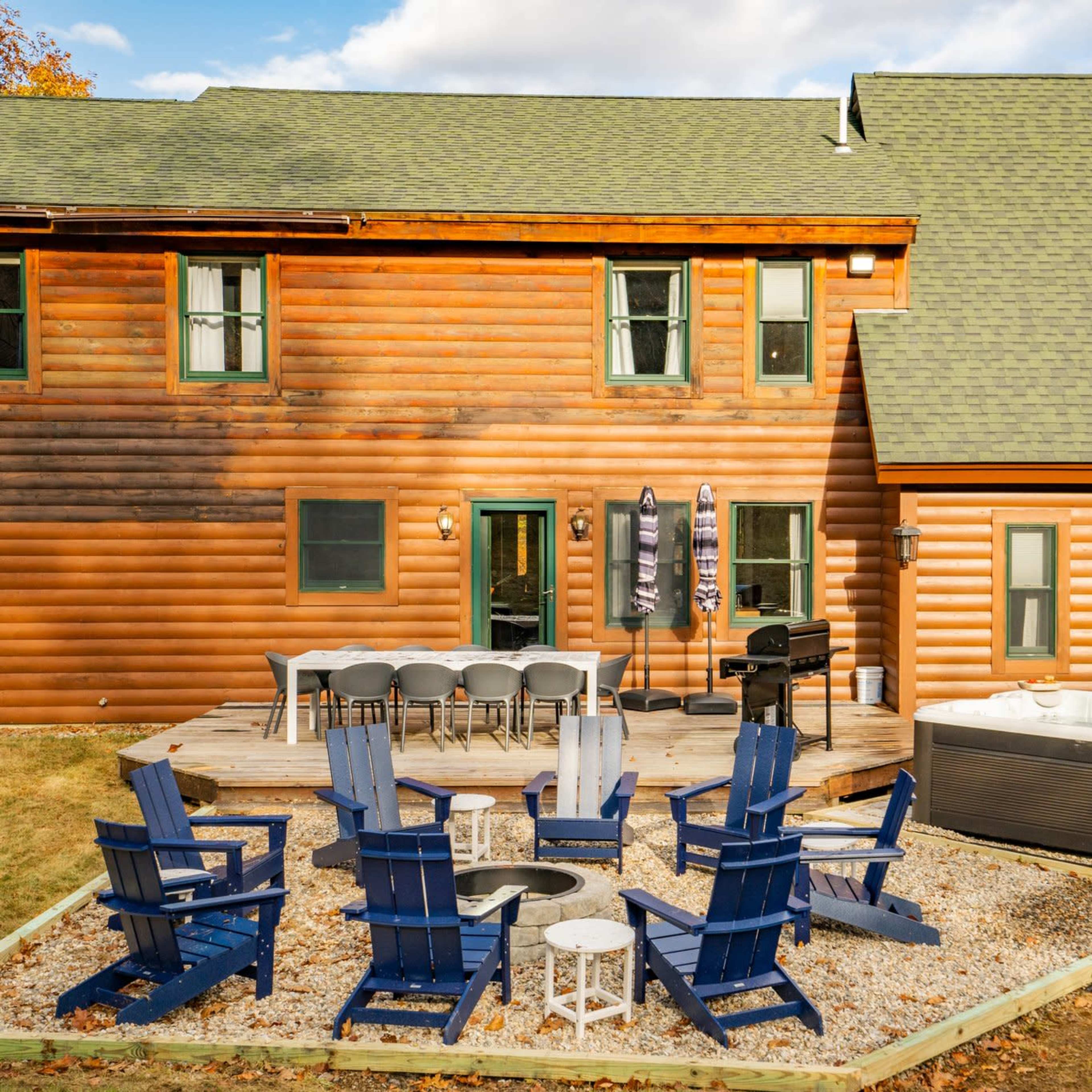 A wooden cabin features a patio with a seating area, outdoor dining table, grill, and a hot tub in a grassy yard.