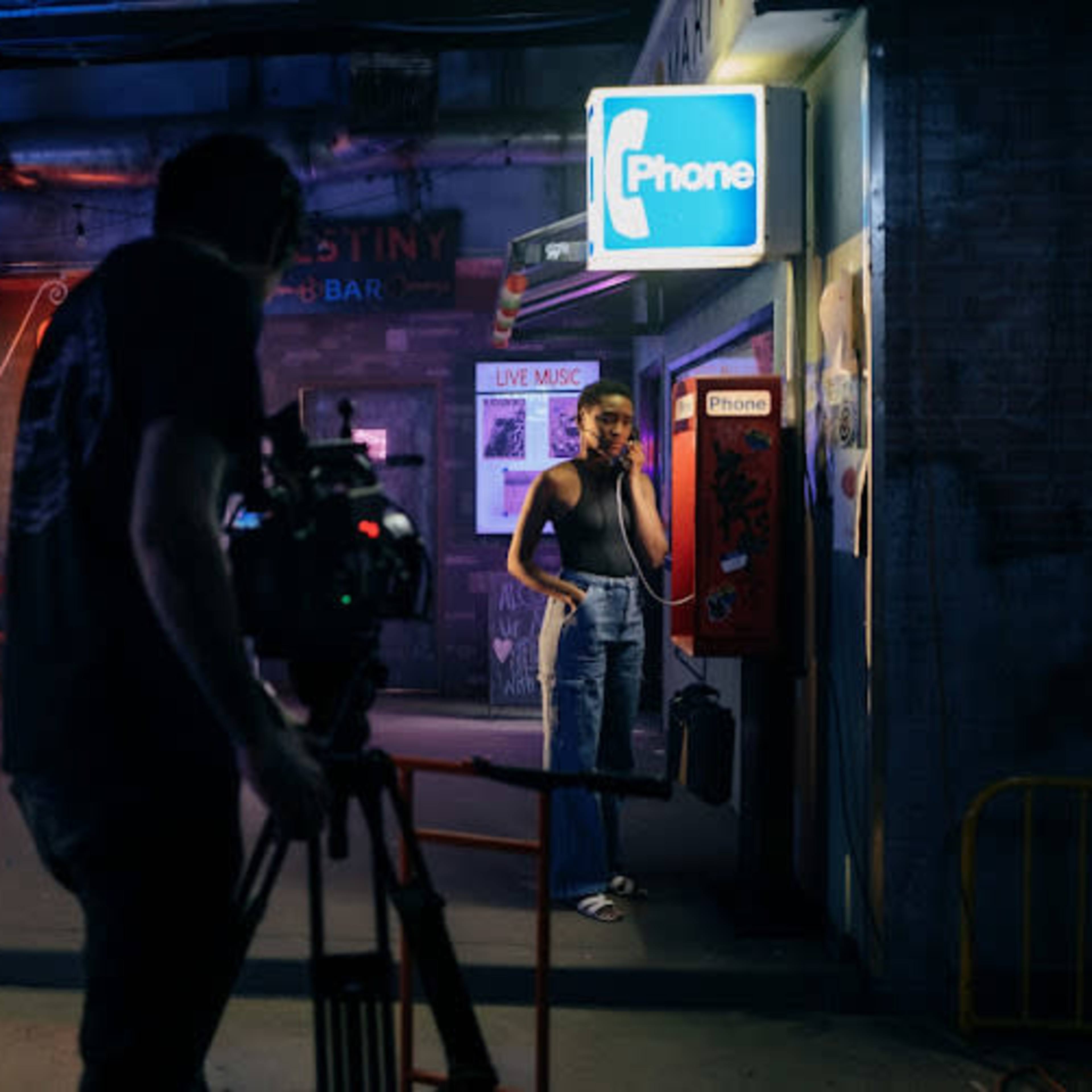 A person stands at a payphone while a cameraman films in a dimly lit urban setting featuring food stalls.