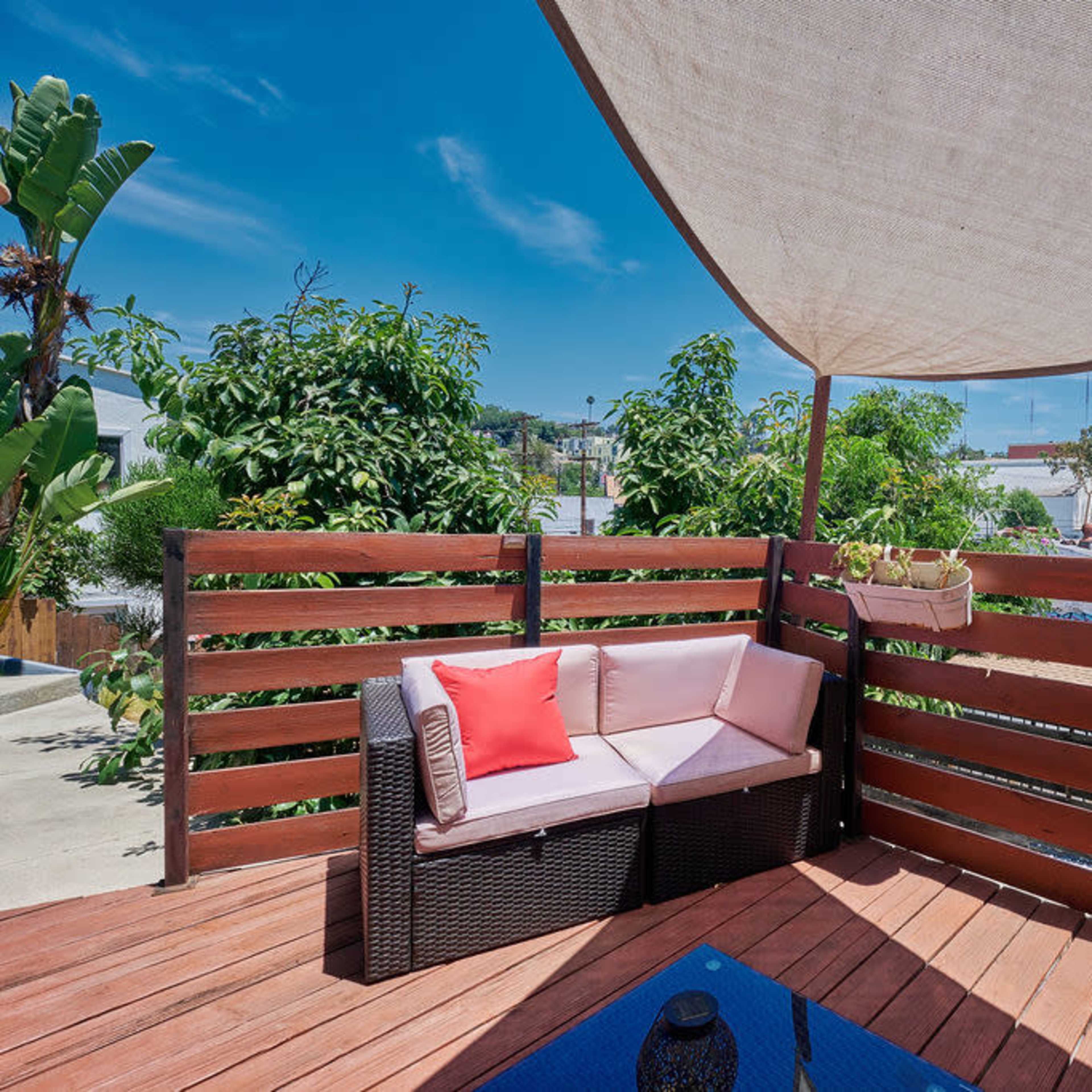 A shaded outdoor seating area with a woven couch, a coffee table, and a view of nearby trees and buildings.