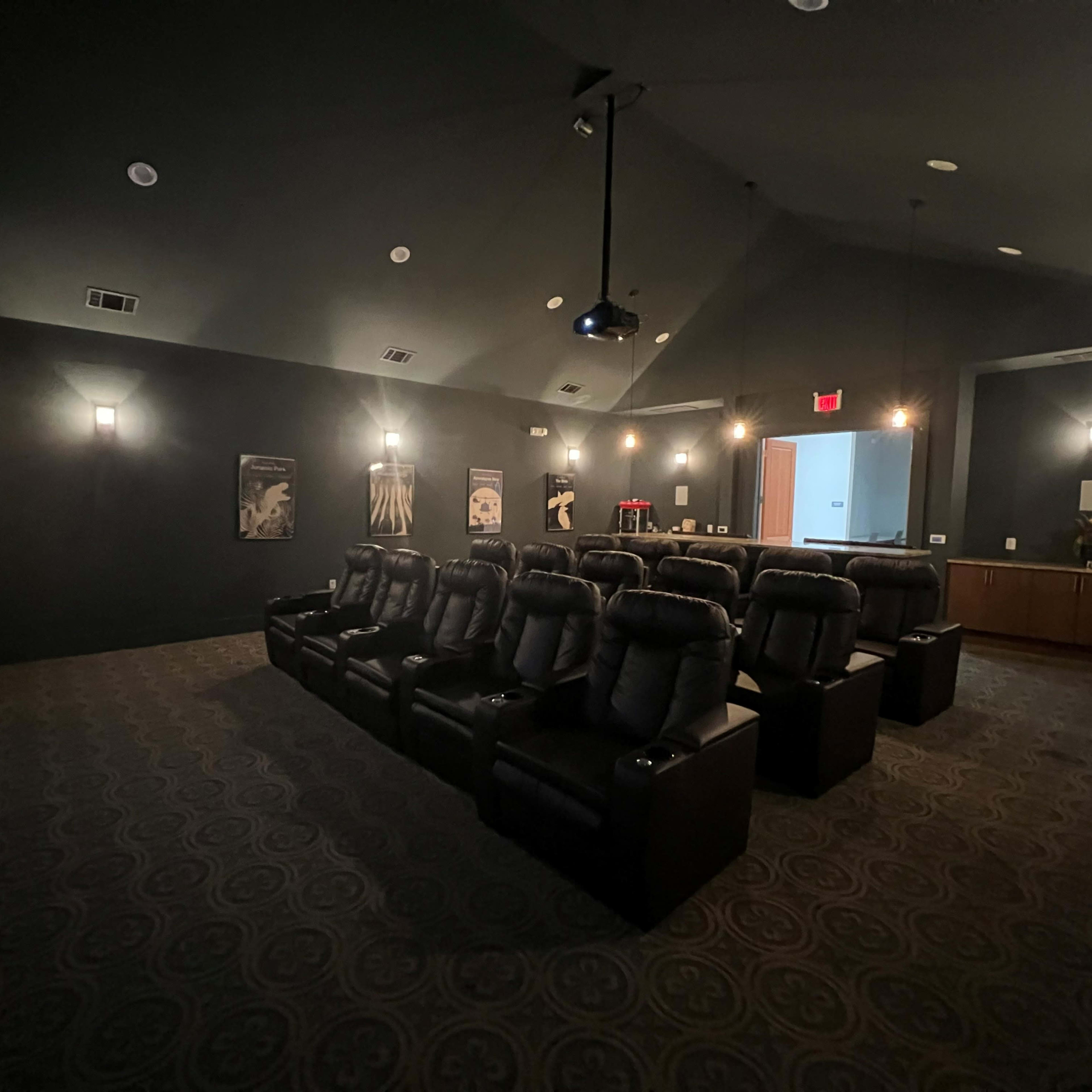 A dark home theater room featuring several black leather recliners arranged in front of a large screen, with dim wall lighting and movie posters on the walls.