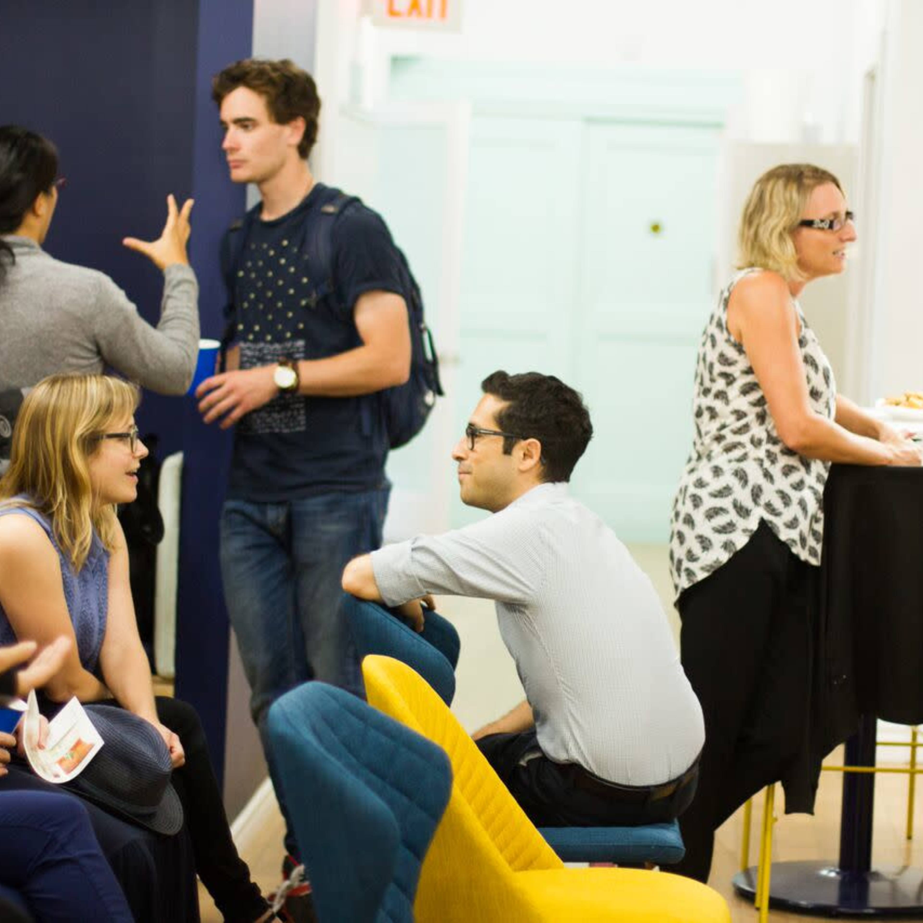 A group of people gathers in a brightly lit room with colorful seating, some engaged in conversation and others seated together.