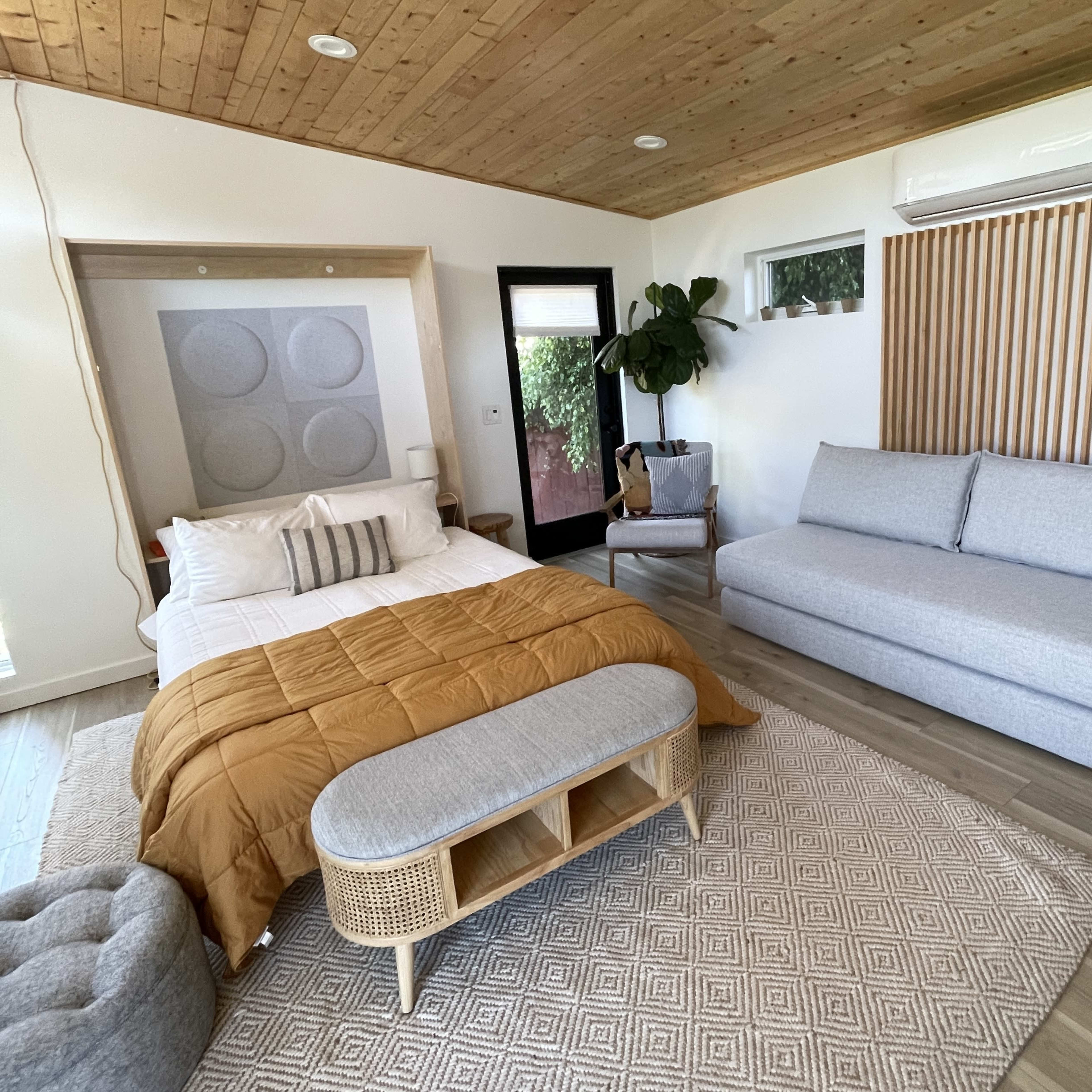 A cozy bedroom featuring a bed with a mustard-colored duvet, a gray sofa, an area rug, and a potted plant, all set against a wooden ceiling and light walls.