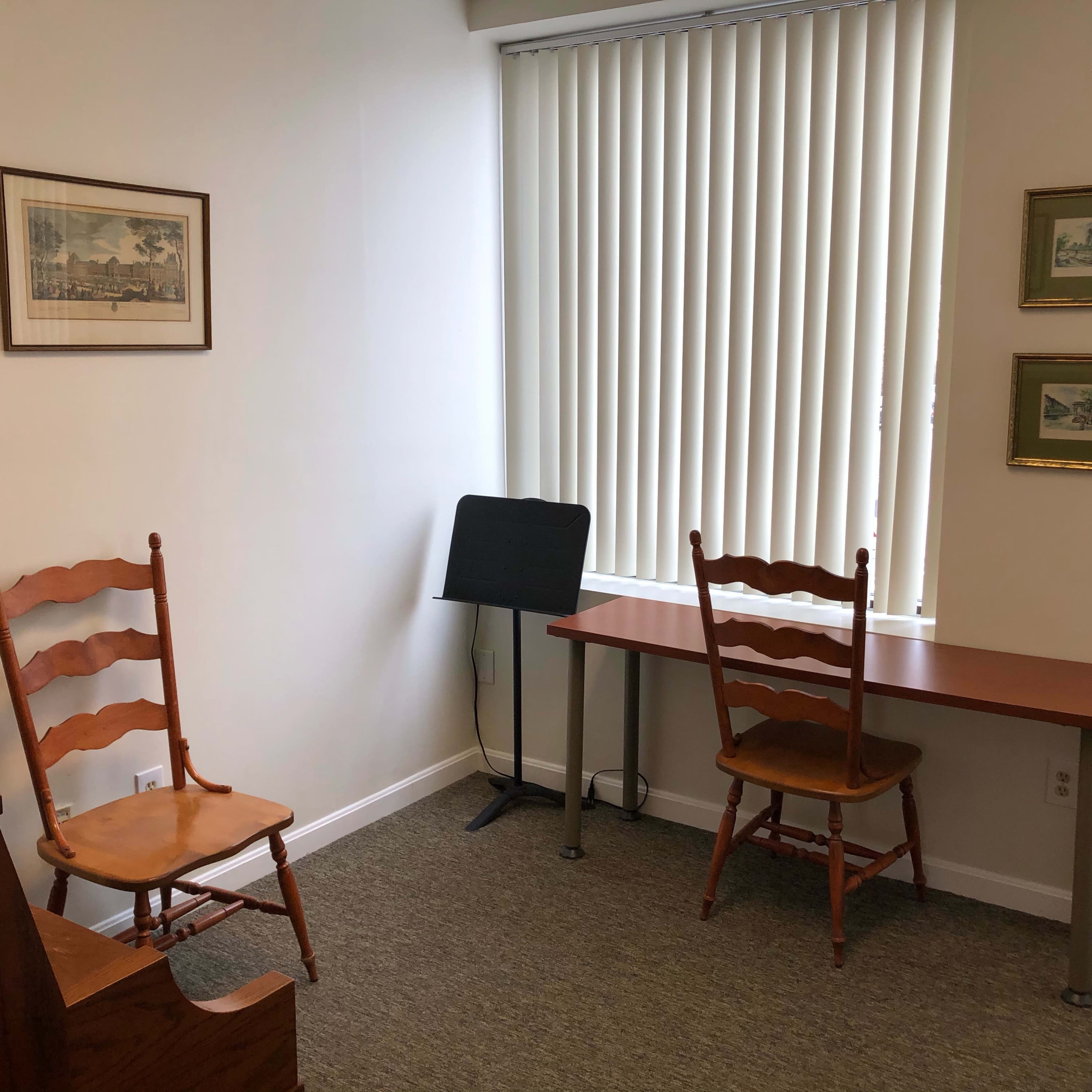 A small room with two wooden chairs, a desk, a music stand, and framed pictures on the wall, illuminated by natural light from a window with vertical blinds.