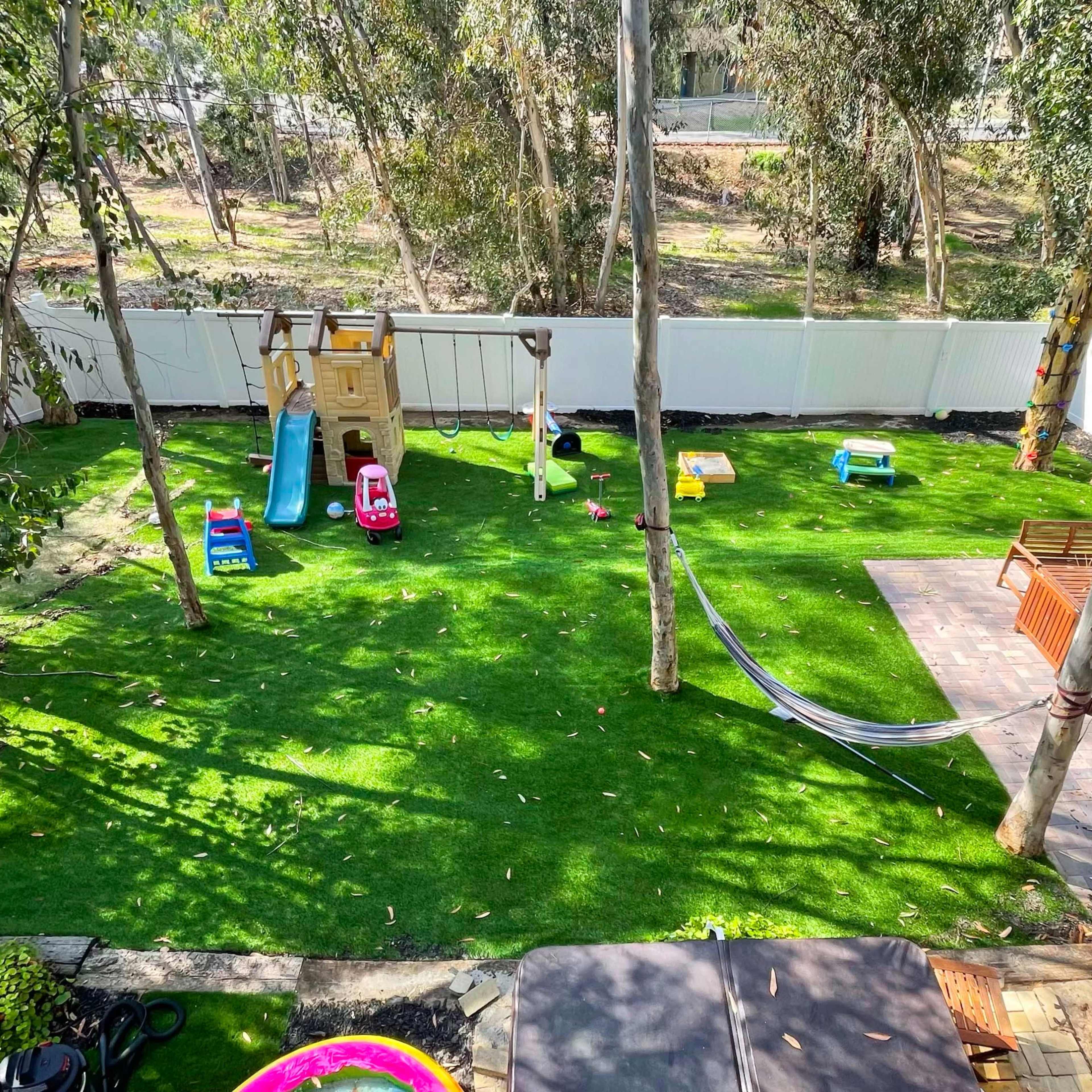 A backyard play area with a playground set, colorful toys, a hammock, and a seating area, all surrounded by trees and a white fence.