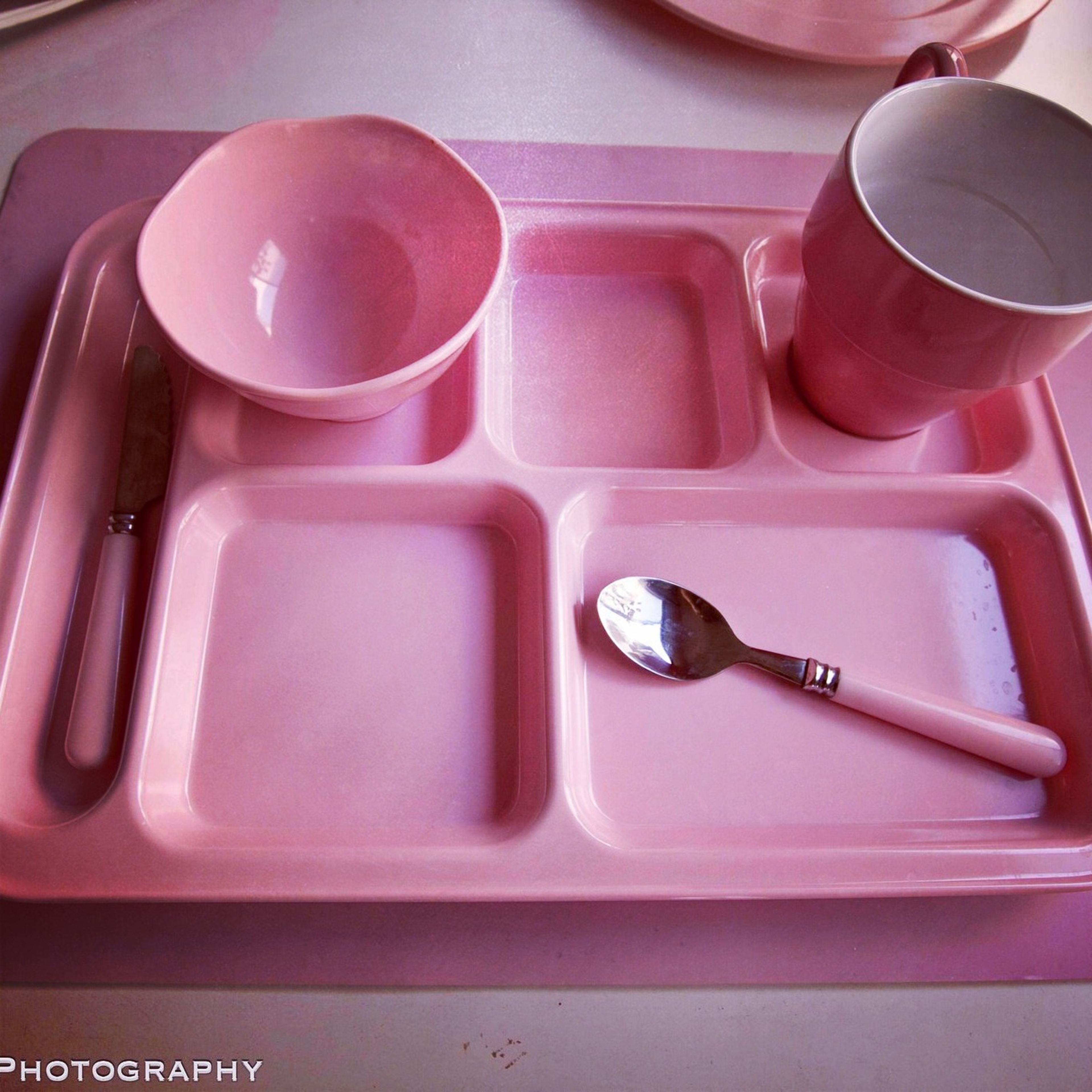 A pink plastic tray with divided sections, accompanied by a pink bowl, mug, knife, and spoon, all arranged on a white surface.