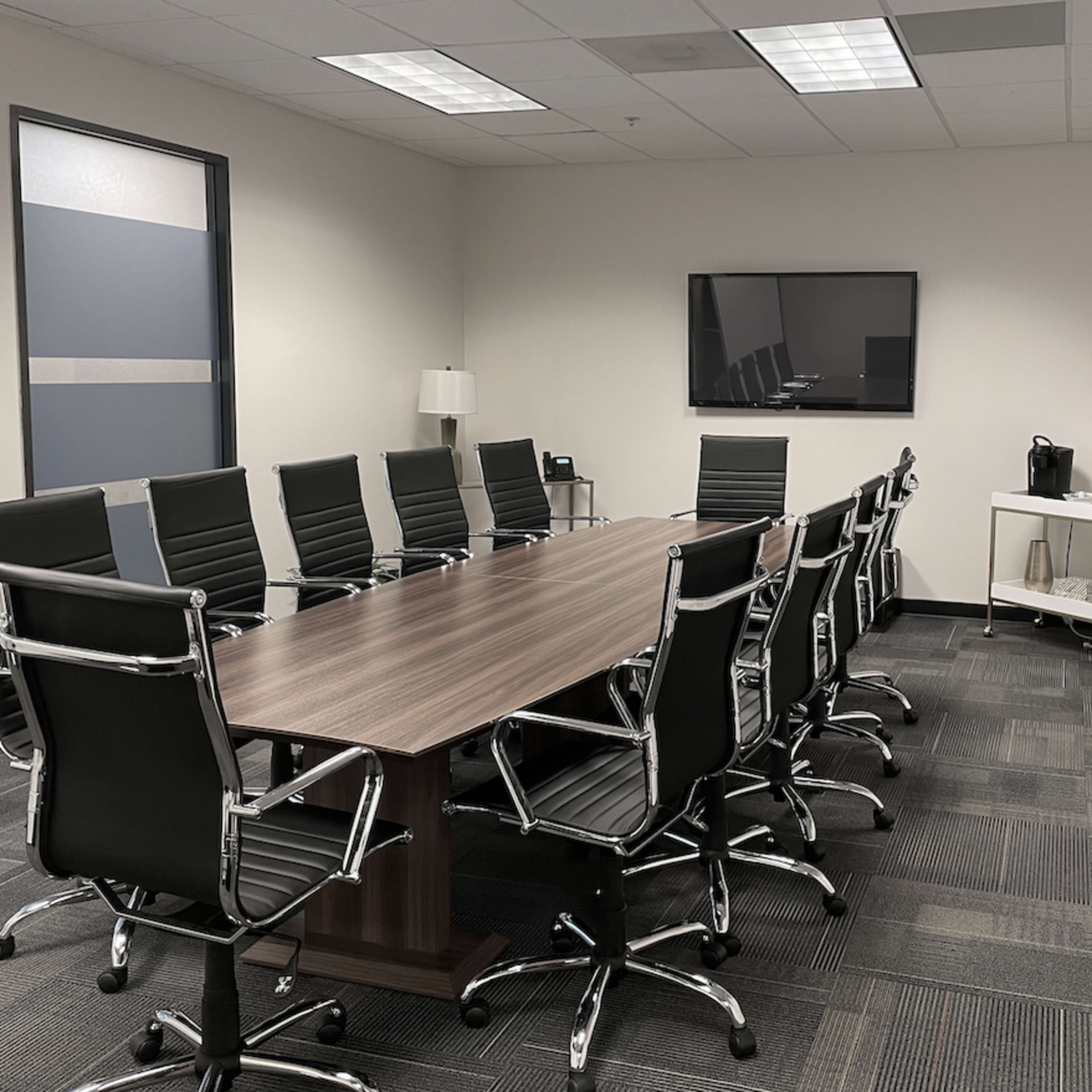 A conference room features a large wooden table surrounded by ten black office chairs, with a television on the wall and a side table holding a coffee maker.