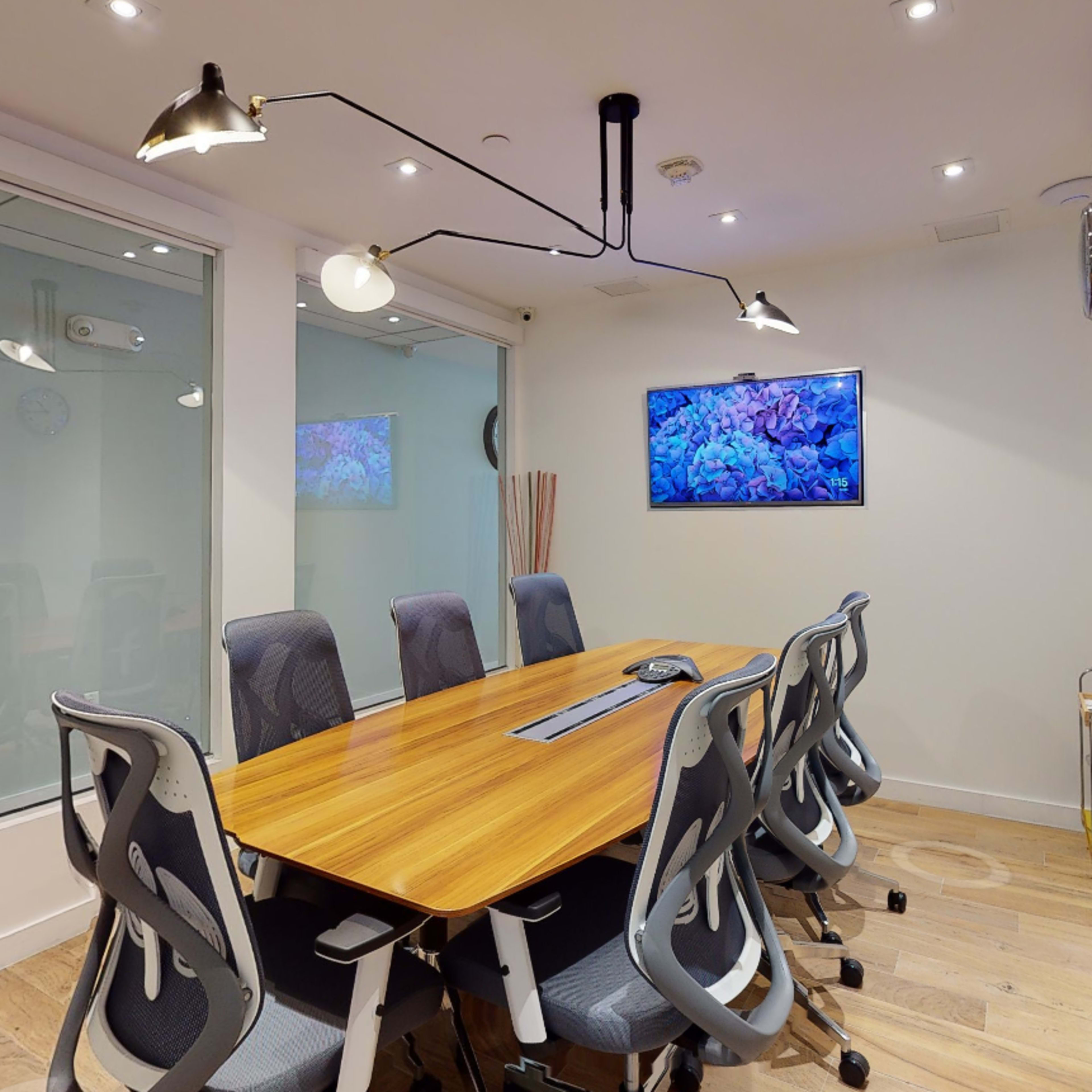 A modern conference room features a long wooden table surrounded by ergonomic chairs, with large glass windows, artistic wall decor, and a digital screen displaying an image.