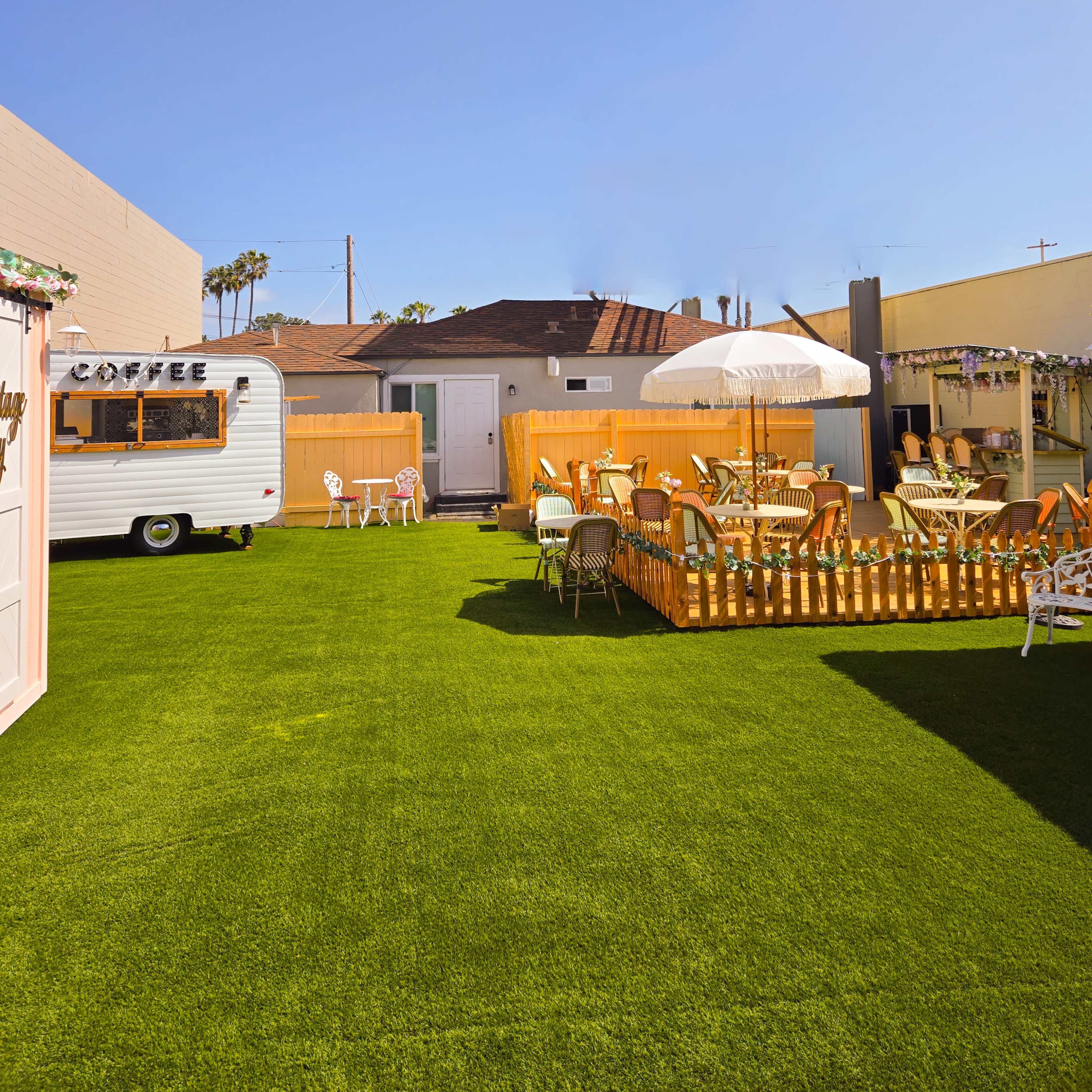 A vibrant, grassy yard with a vintage camper, outdoor seating, and a wooden fence under a clear blue sky.
