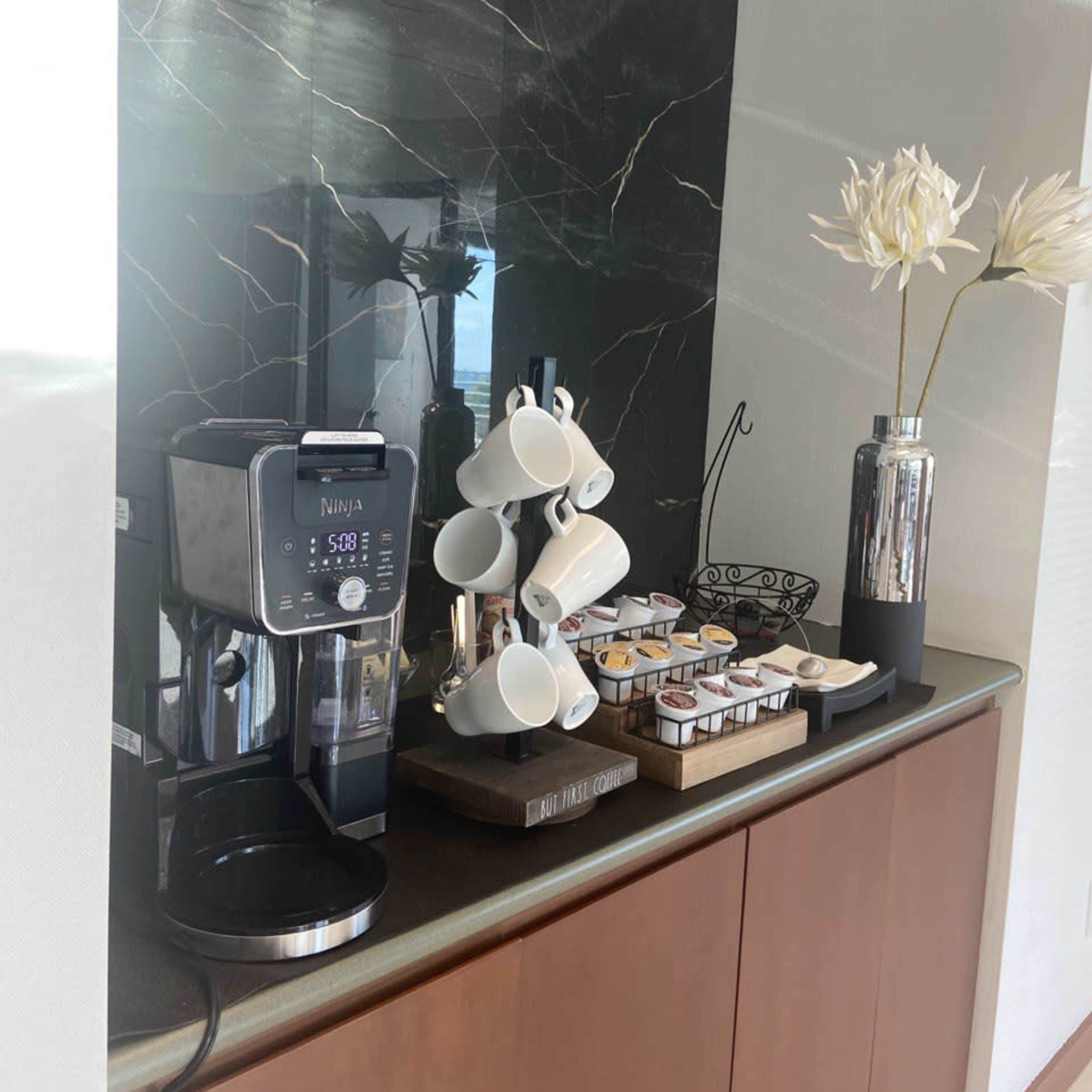 A kitchen counter with a Ninja coffee maker, a tiered rack of coffee pods, a holder for cups, a decorative vase with flowers, and a dark marble backsplash.