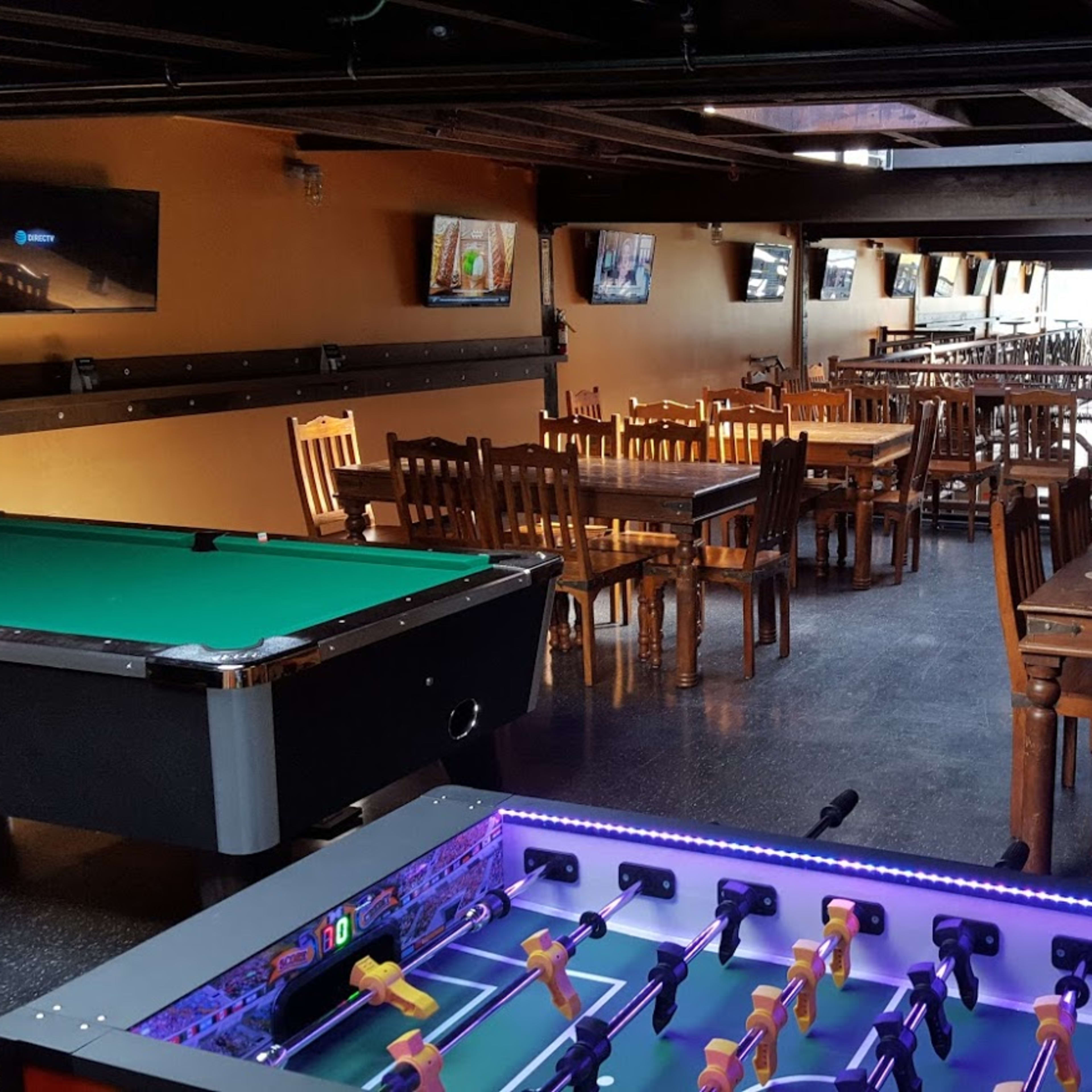 A spacious indoor venue featuring a pool table, foosball table, and several wooden dining tables with chairs.