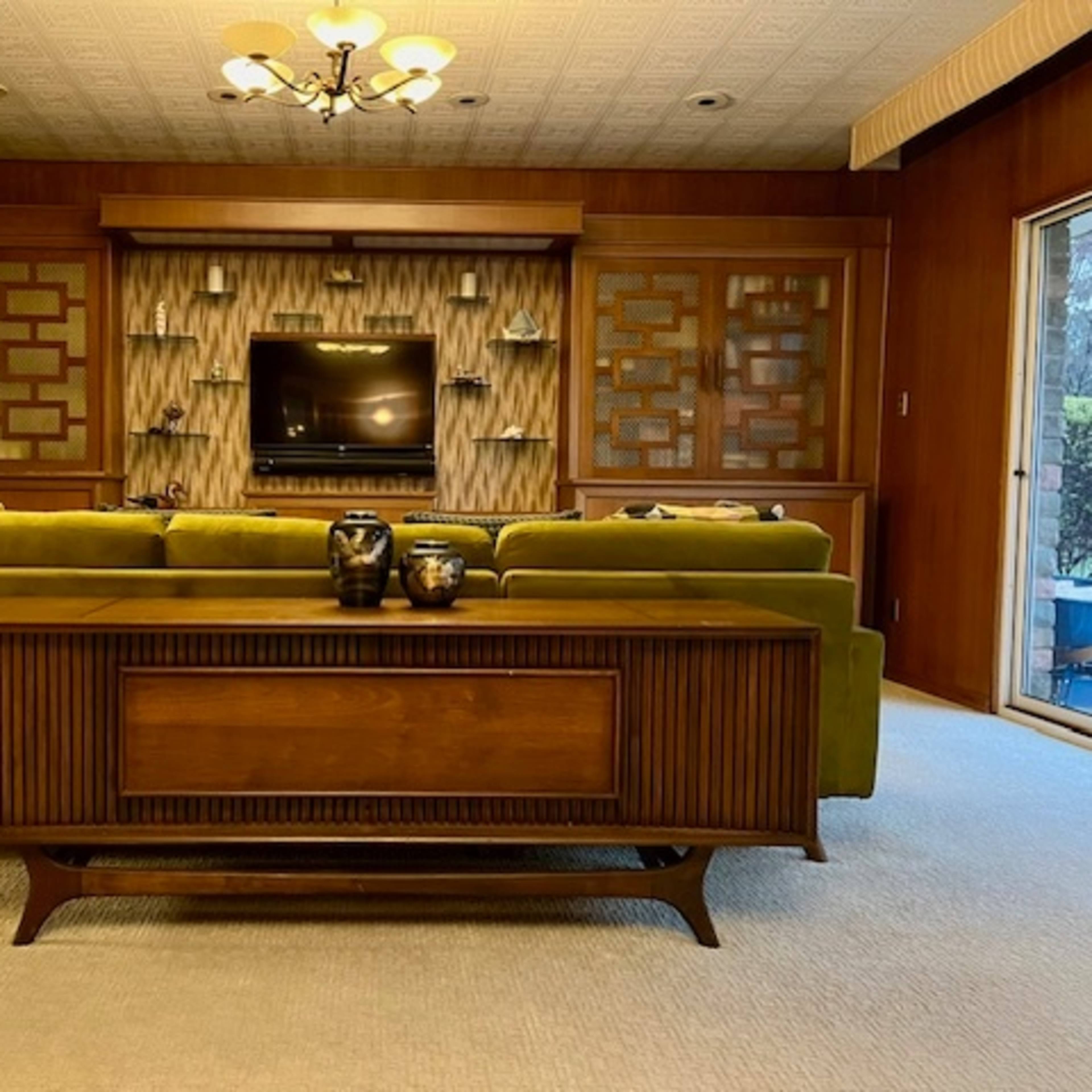 A living room with a green couch facing a wall-mounted television, surrounded by wooden cabinetry and large windows.