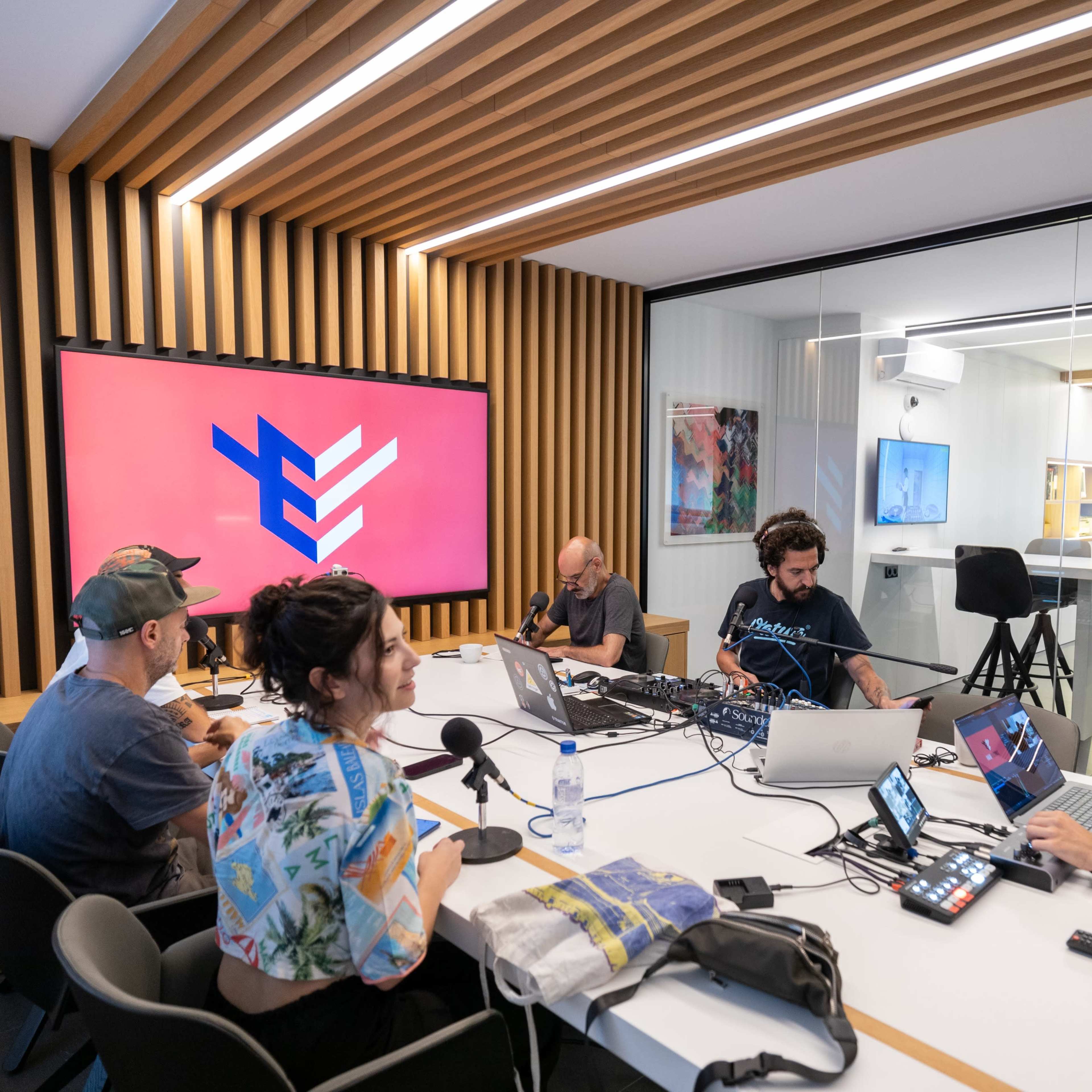 A group of five people collaborates around a white table, engaging with laptops and microphones in a modern workspace featuring a large screen displaying colorful graphics.