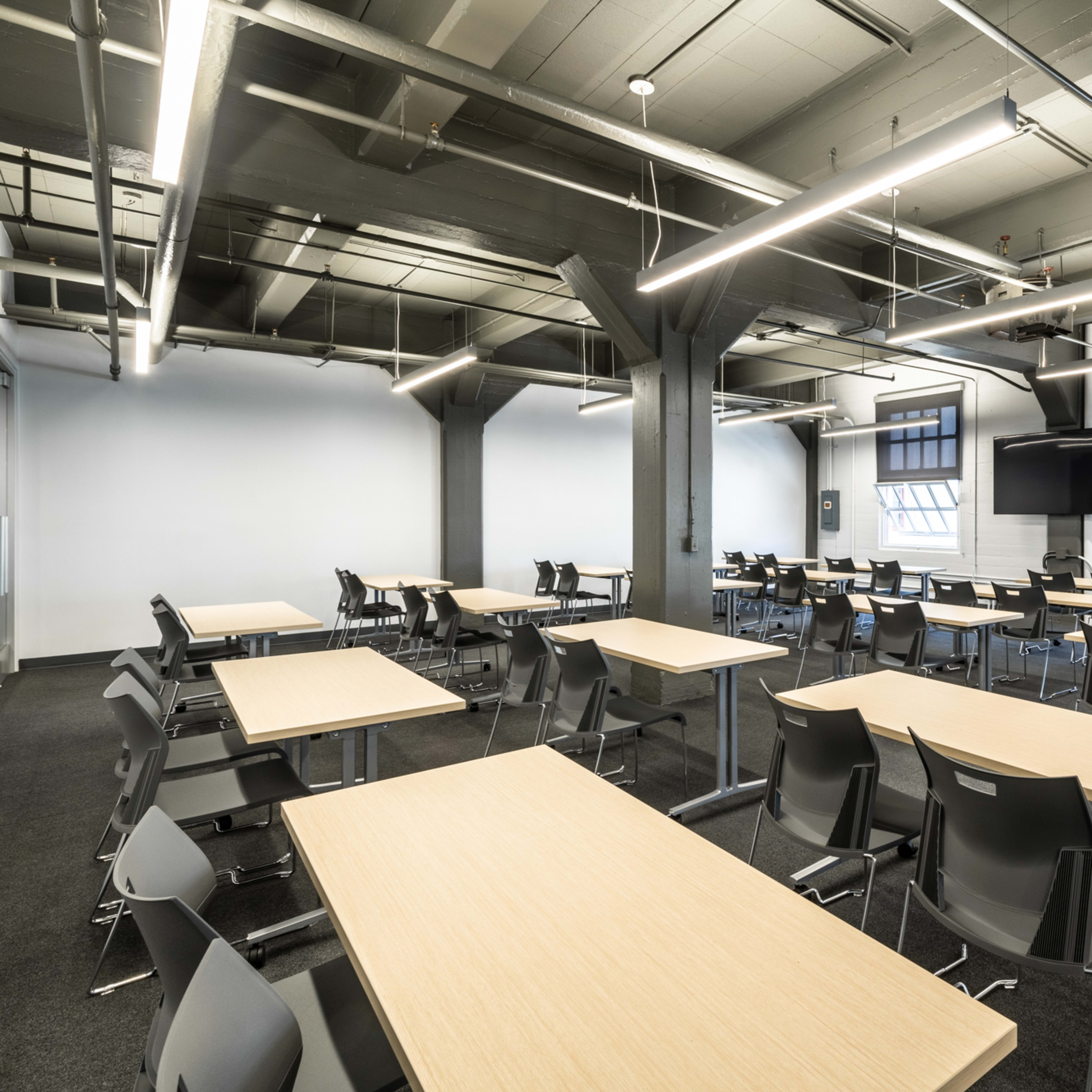 A spacious classroom features several rows of wooden tables and black chairs under industrial-style lighting.