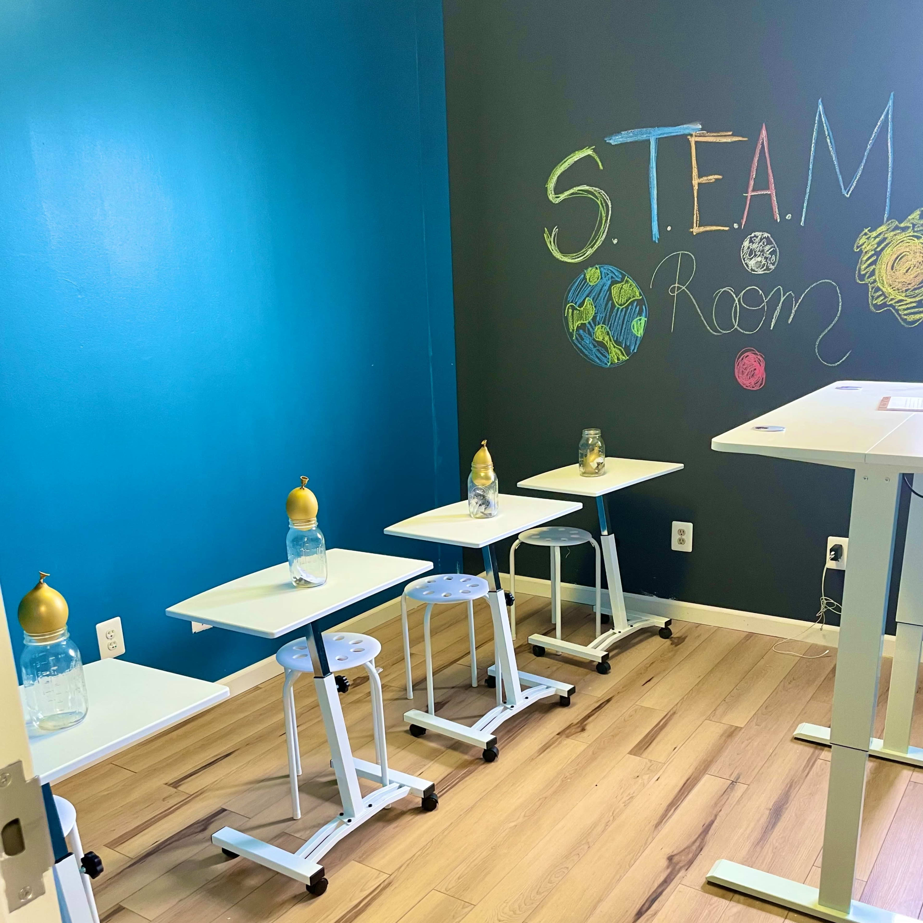 A small classroom with four movable desks, each featuring a water bottle, and a wall painted with "S.T.E.A.M. Room."