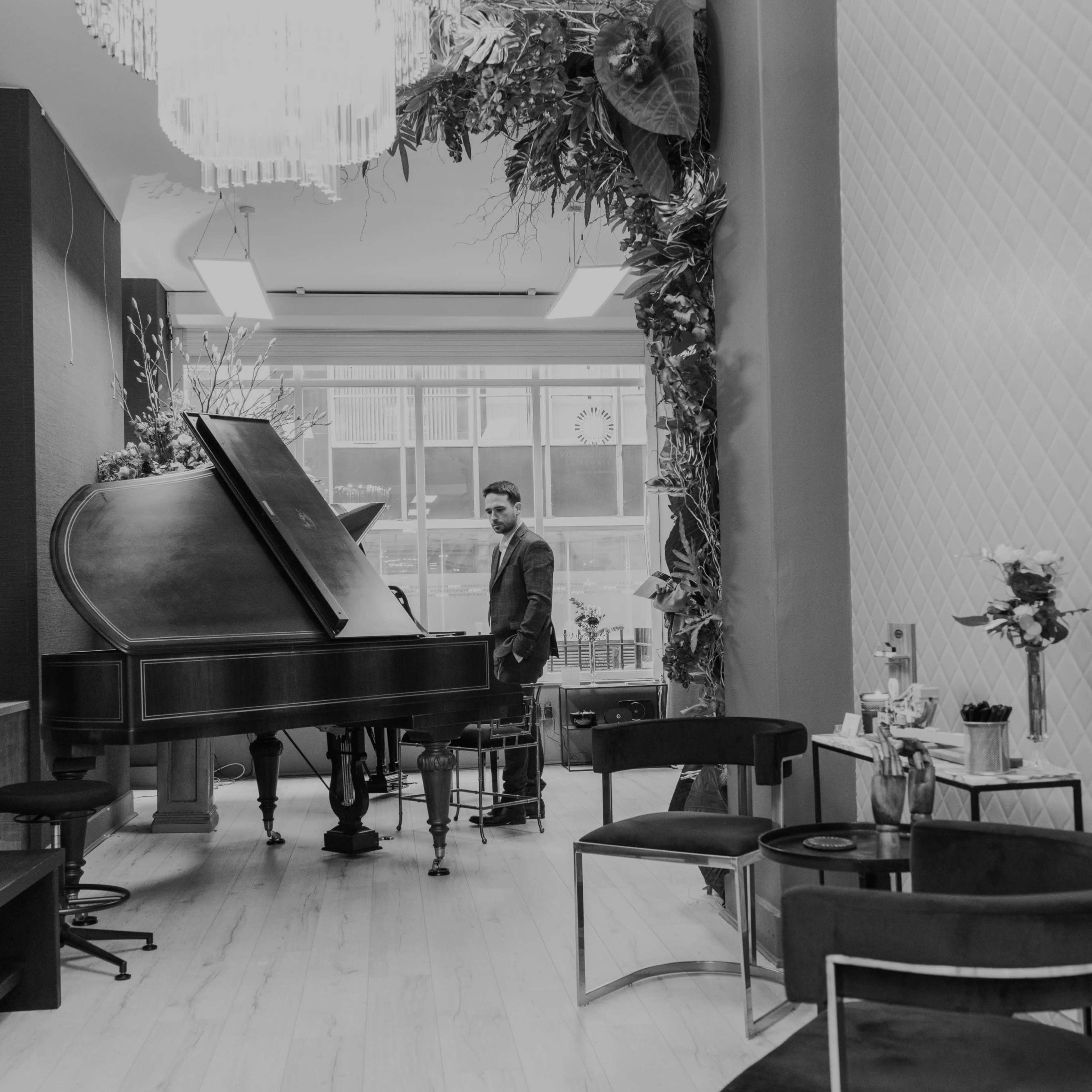 A man stands beside a grand piano in a modern, plant-decorated room with stylish seating and a chandelier.