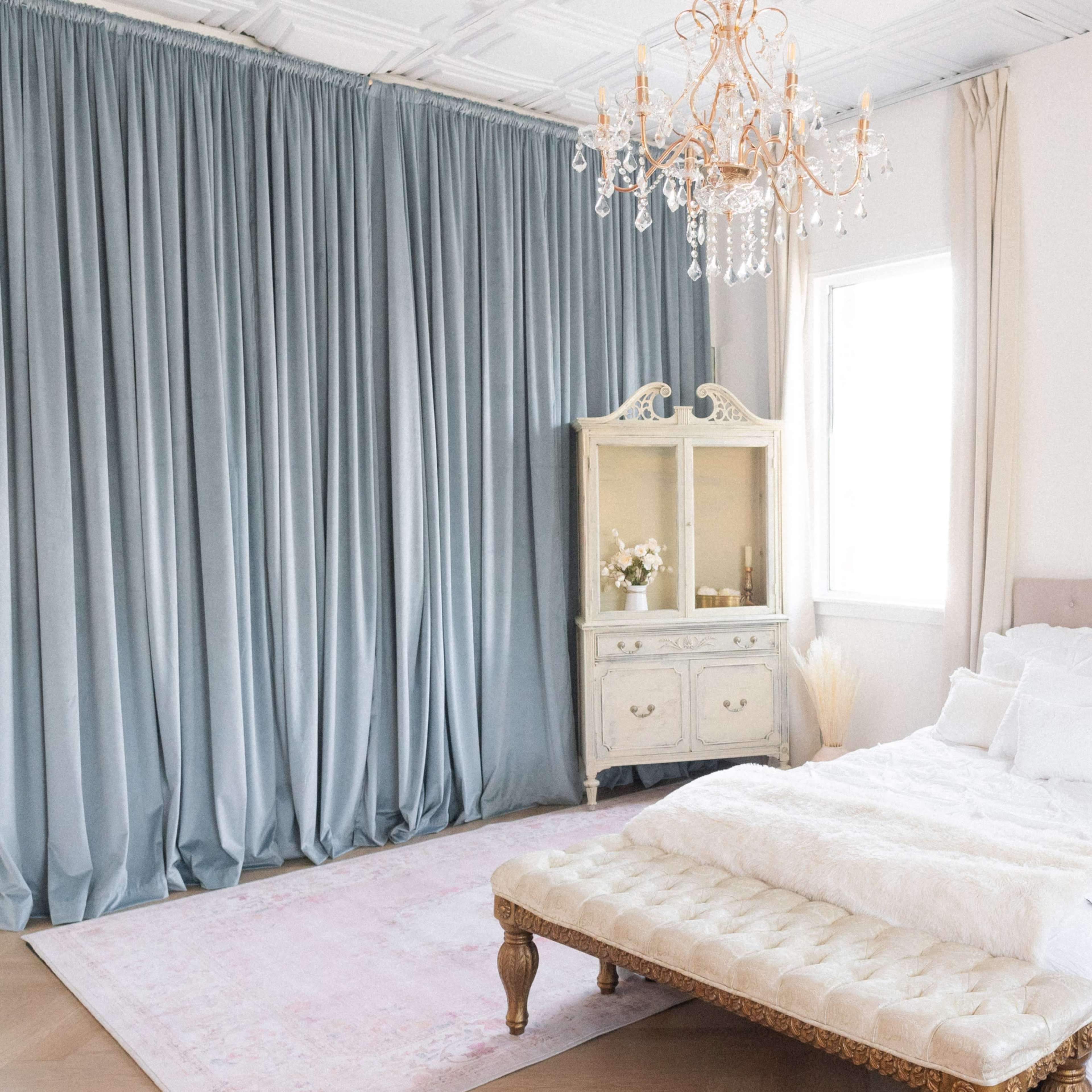A bright bedroom features a bed with white bedding, a tufted bench, a vintage-style cabinet, and elegant curtains framing the windows.