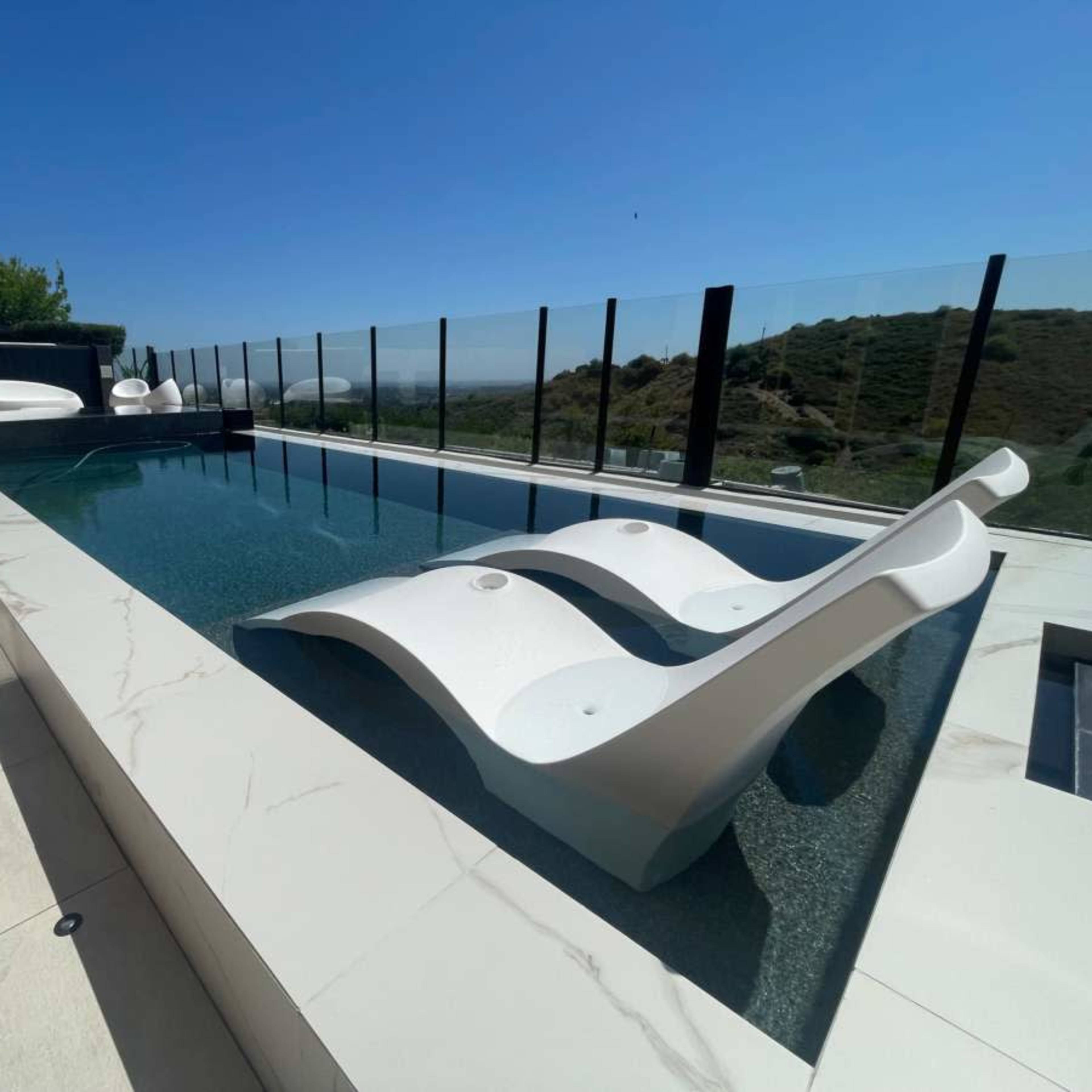 A modern swimming pool with white lounge chairs and a glass railing overlooking a scenic landscape.