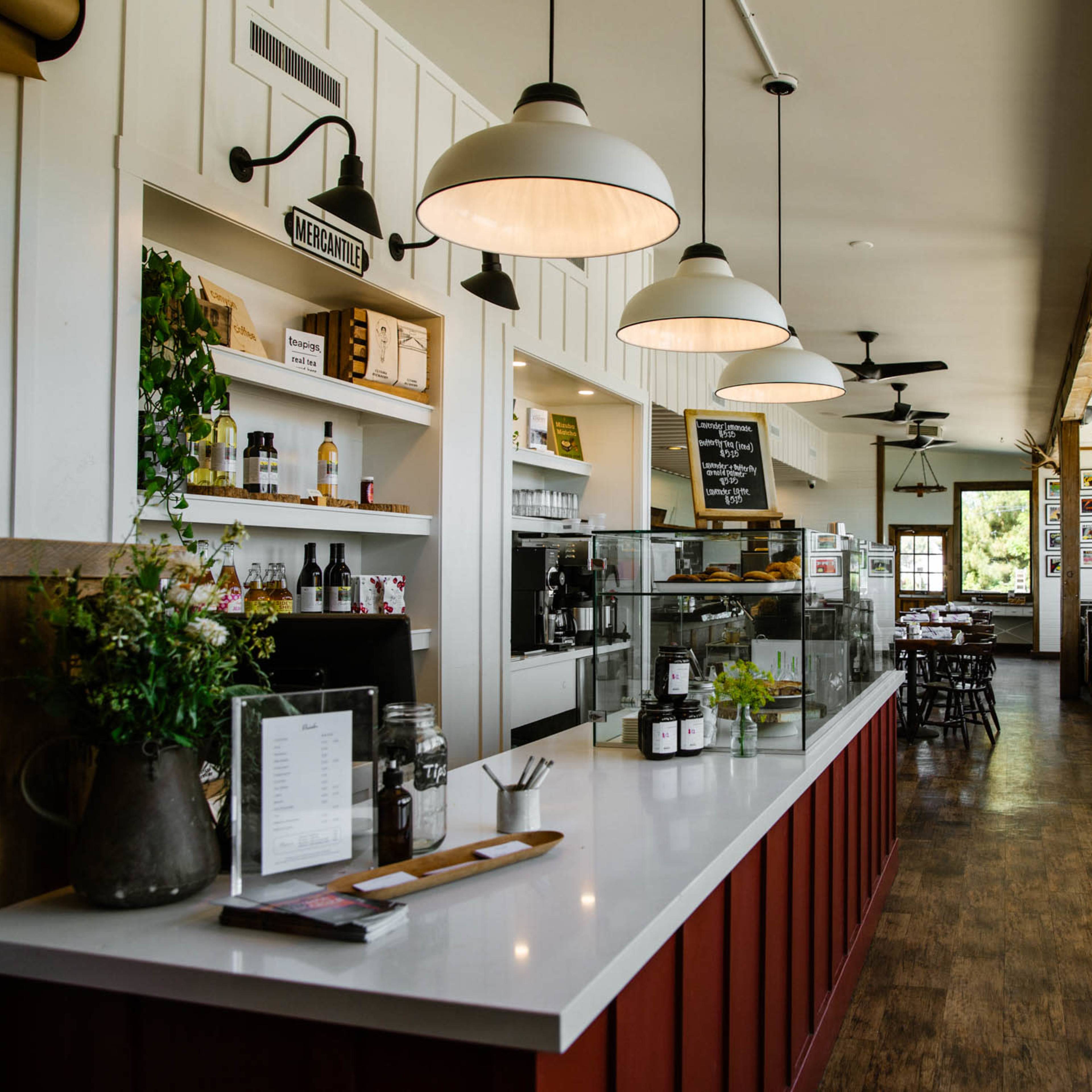 A cafe interior featuring a service counter with a display case, shelves of products, and tables along large windows.