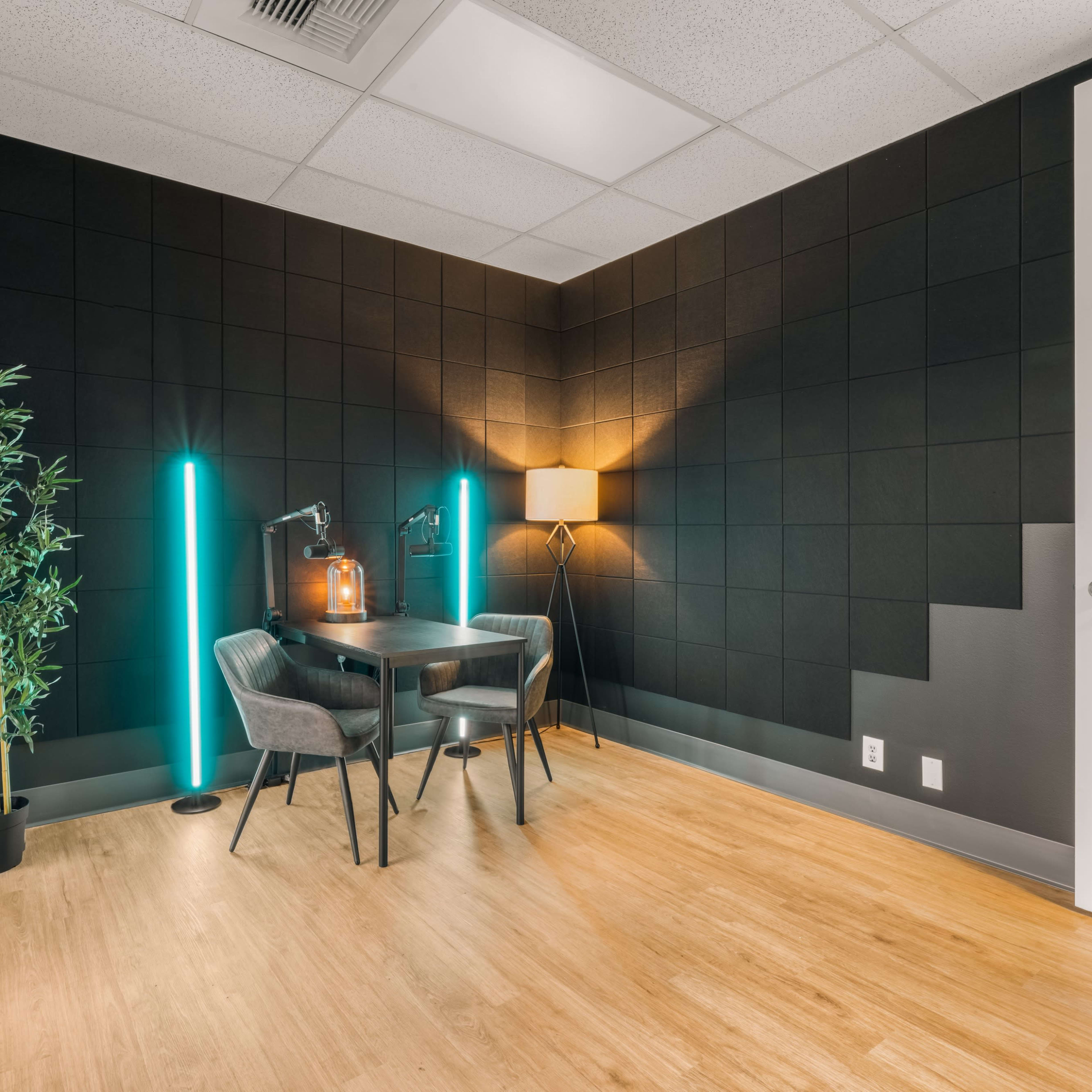 A small office space features a black wall, a wooden floor, a desk with two chairs, and lighting from a lamp and two standing lights.