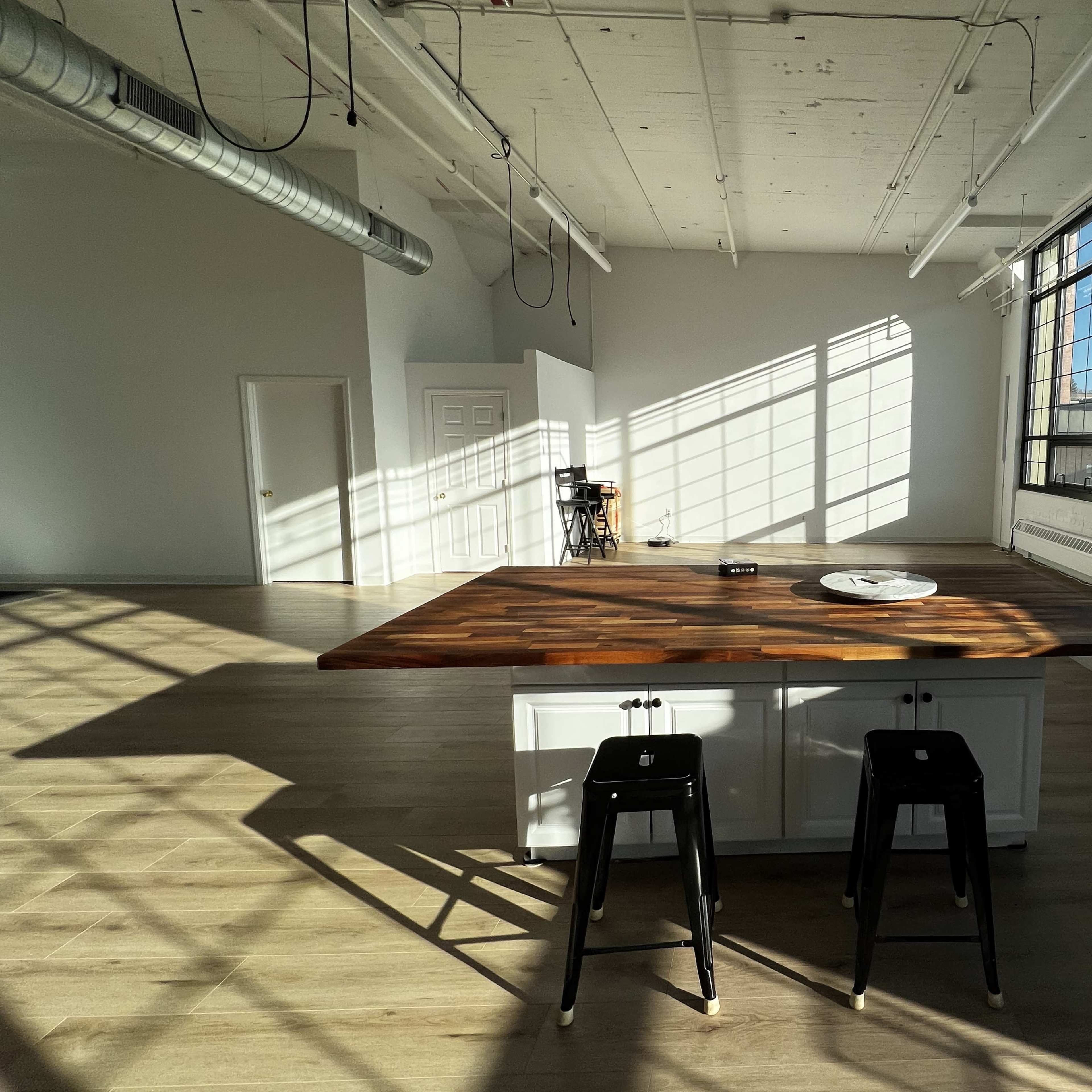 A spacious, sunlit room features a large wooden island with black stools and long shadows cast by sunlight through large windows.