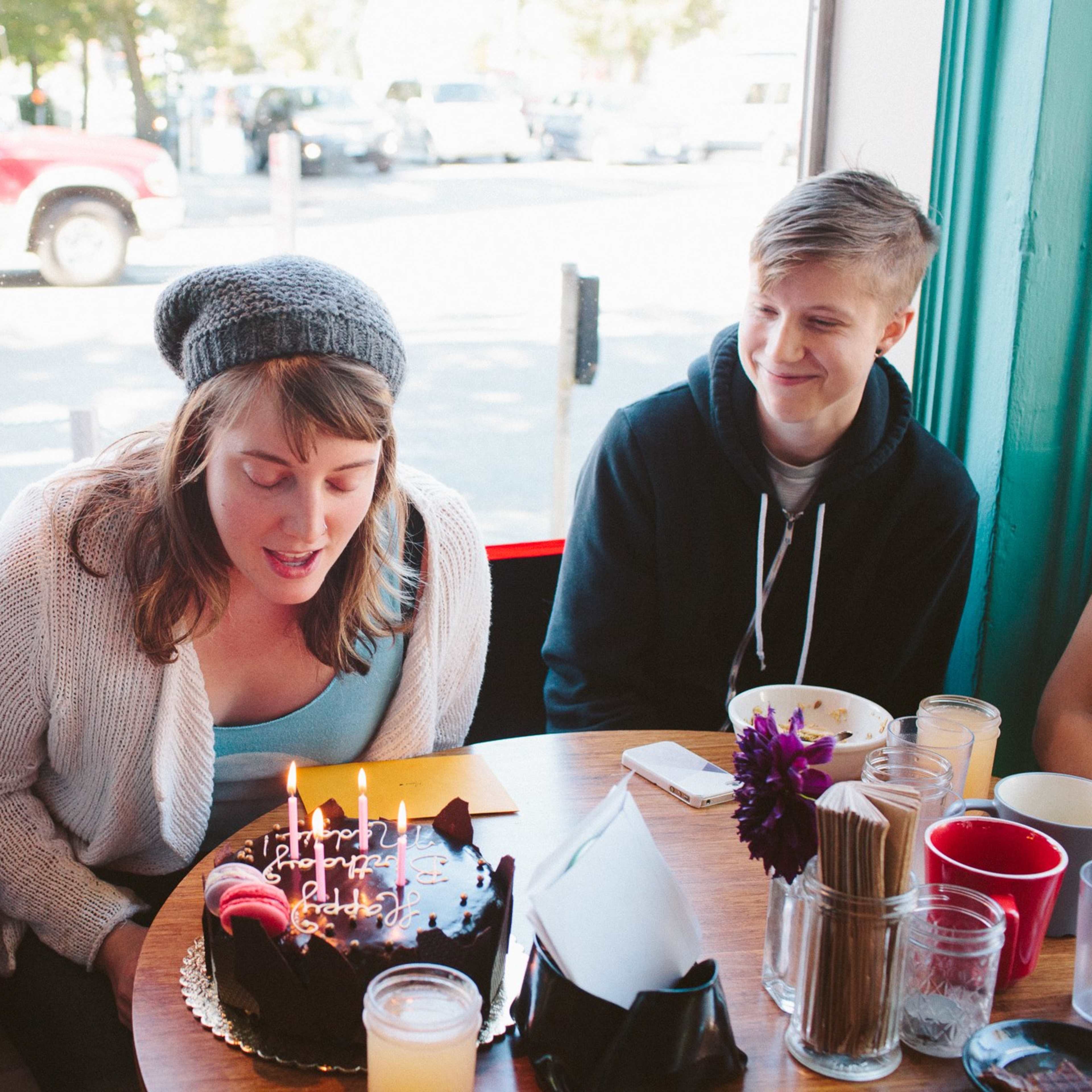 A group of friends gathers around a table with a birthday cake as one person prepares to blow out the candles.