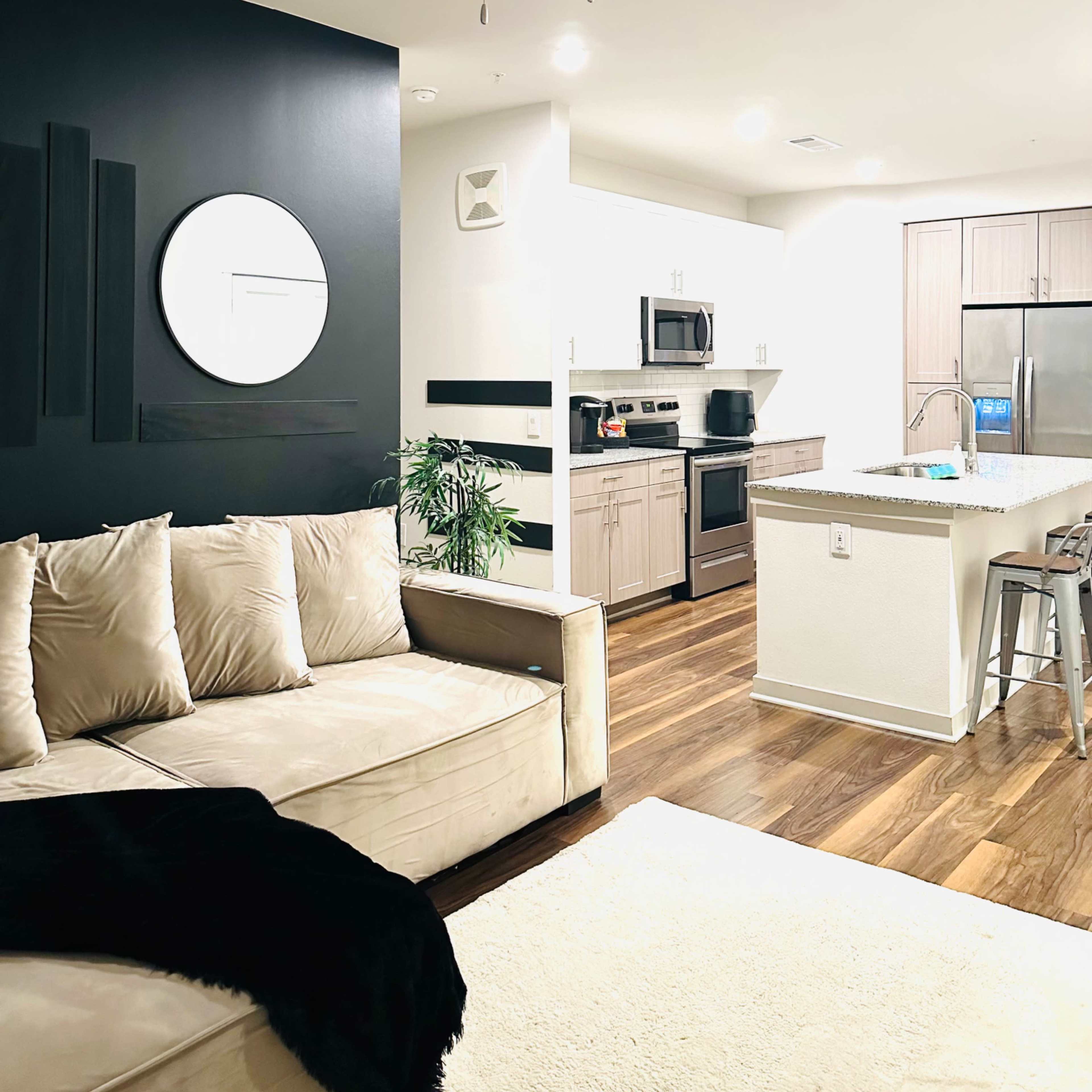 A modern living room and kitchen area featuring a beige sectional sofa, a wall-mounted round mirror, and stainless steel appliances.