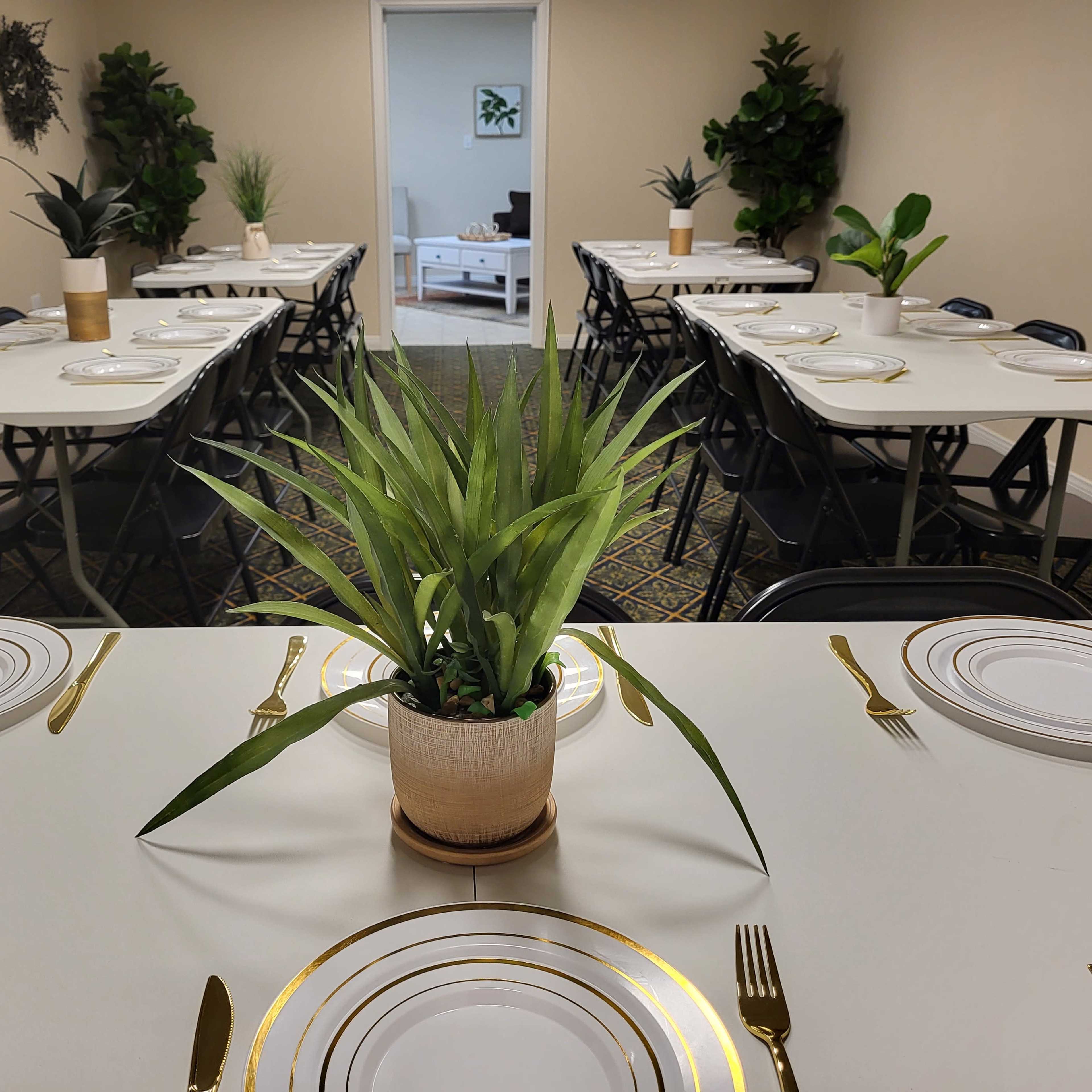 A neatly arranged dining area features several tables set with plates, utensils, and potted plants at the center.