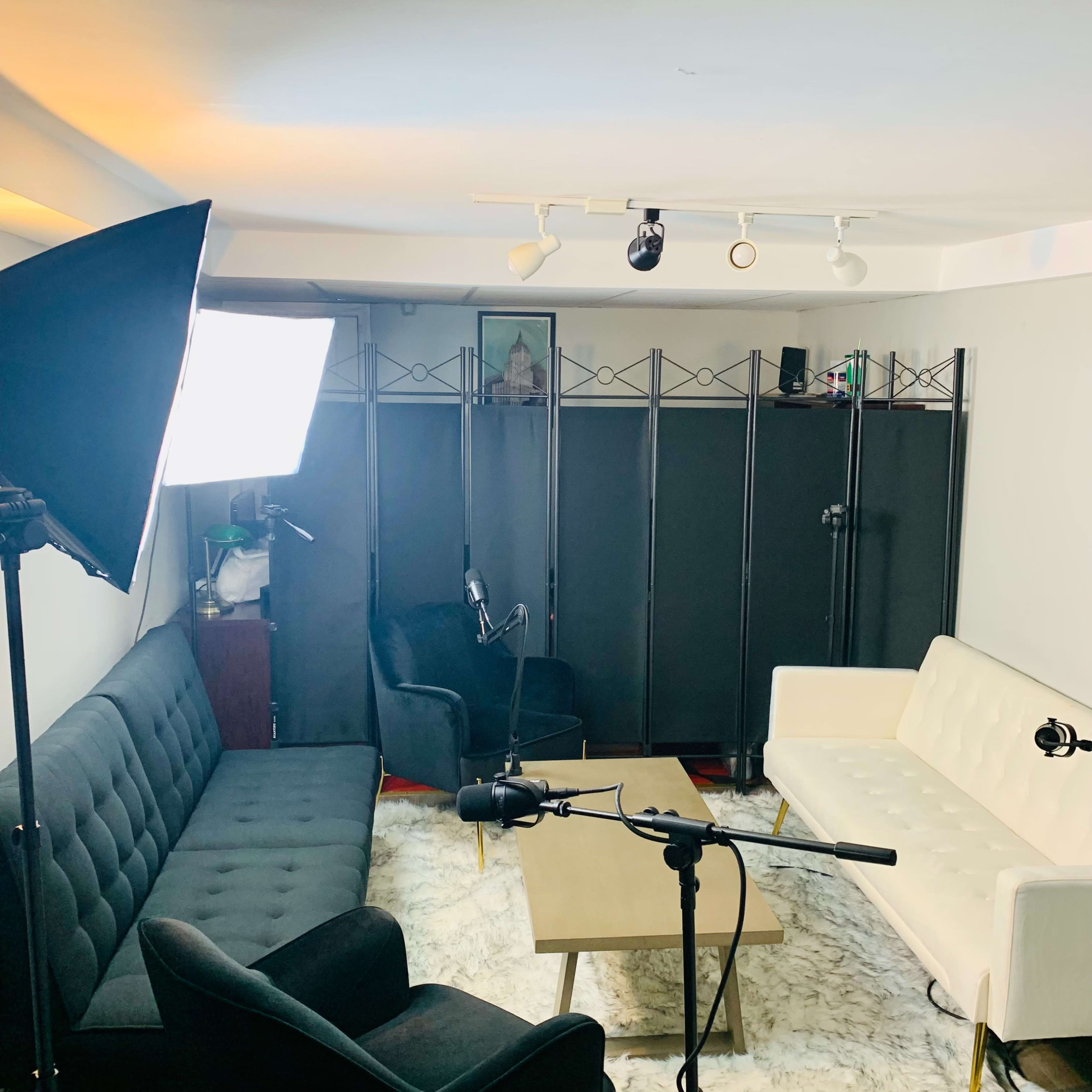 A small, well-lit recording studio with two sofas, a coffee table, and microphones set up in front of a black folding screen.