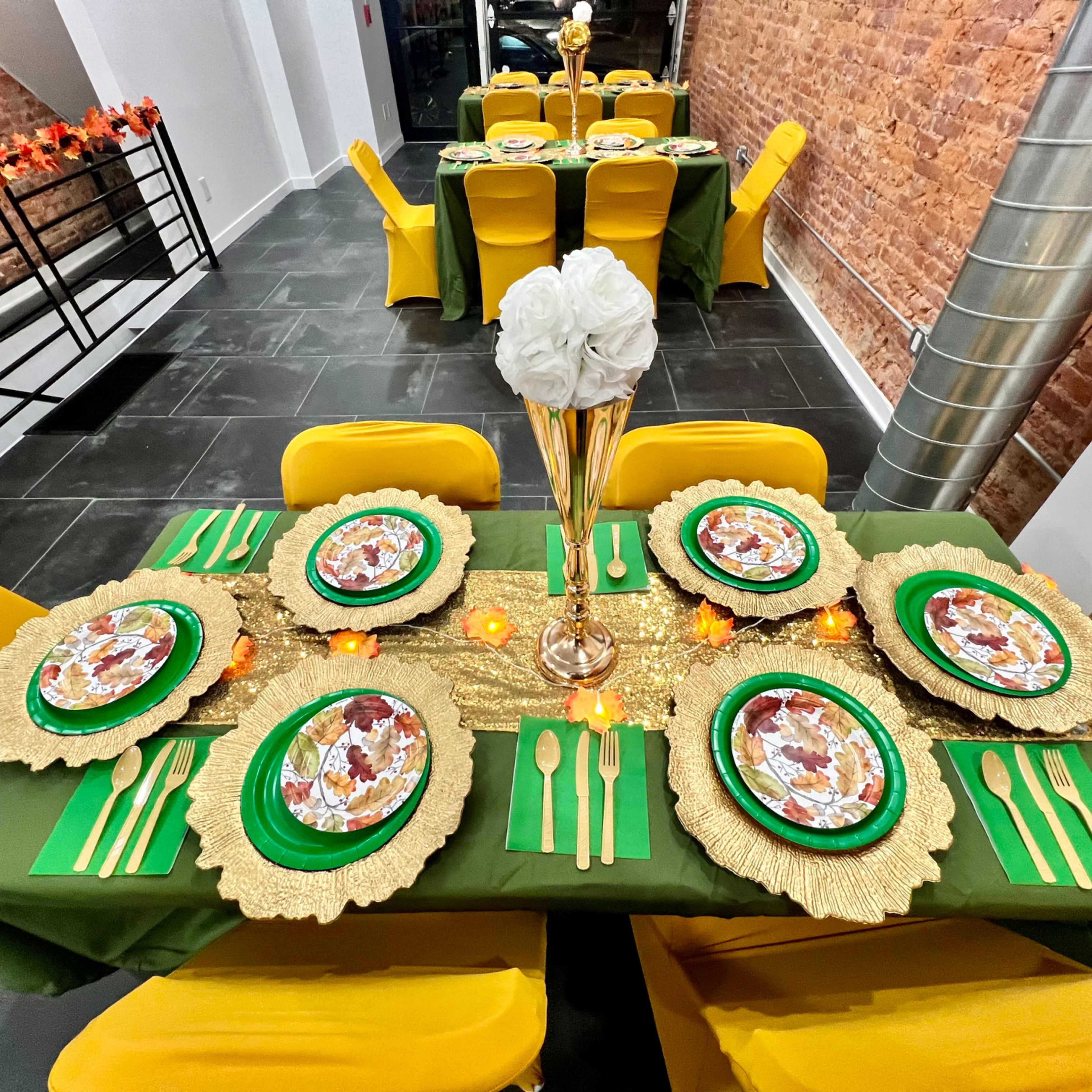 A dining table is set with green and gold decorations, featuring plates with floral designs, utensils, and a tall flower vase amidst a backdrop of a brick wall and yellow chairs.