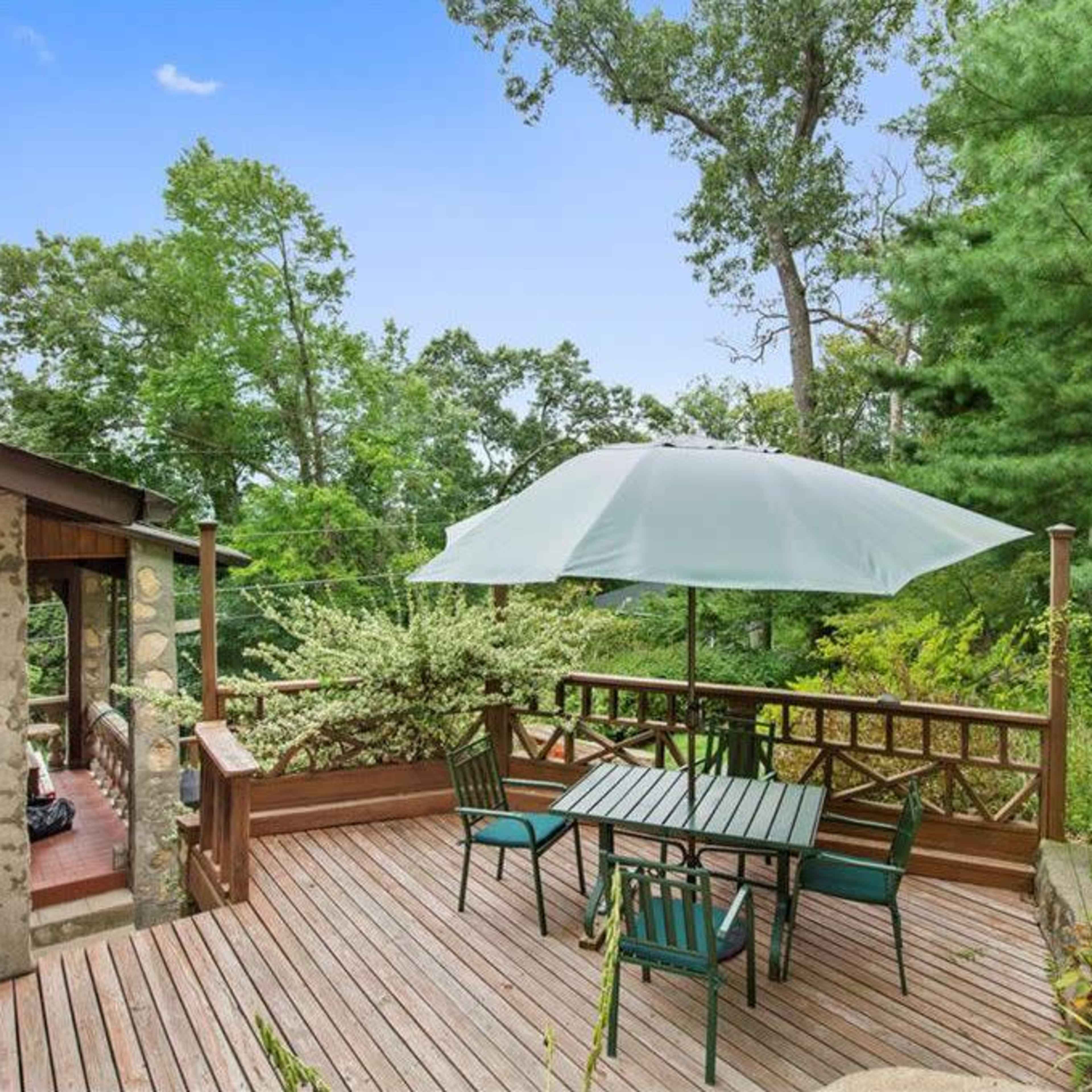 A wooden deck features a table and chairs beneath a large umbrella, surrounded by trees and greenery.