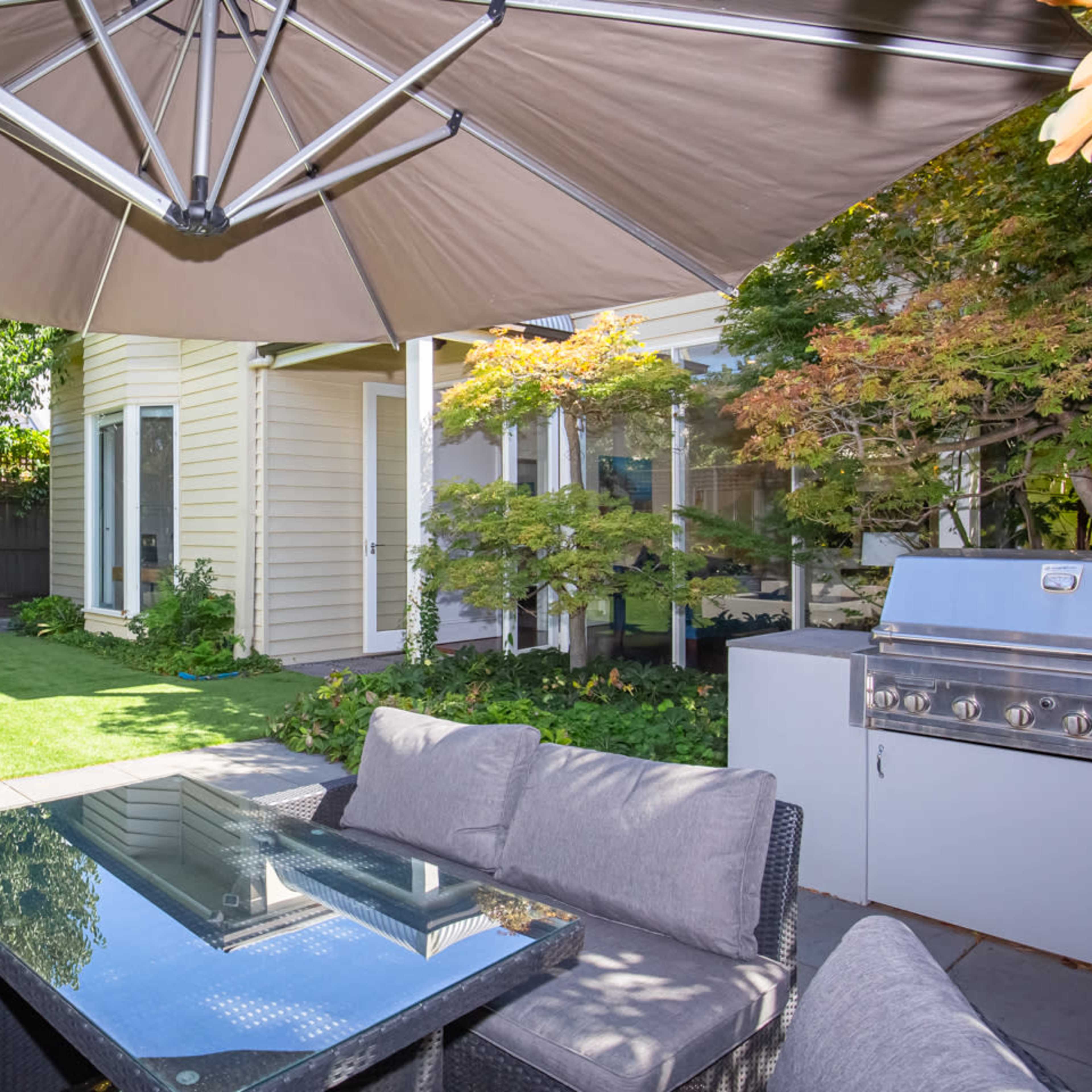 A patio area features a glass-top table surrounded by cushioned seating, with an umbrella above and a grill nearby, adjacent to a landscaped garden and a house.