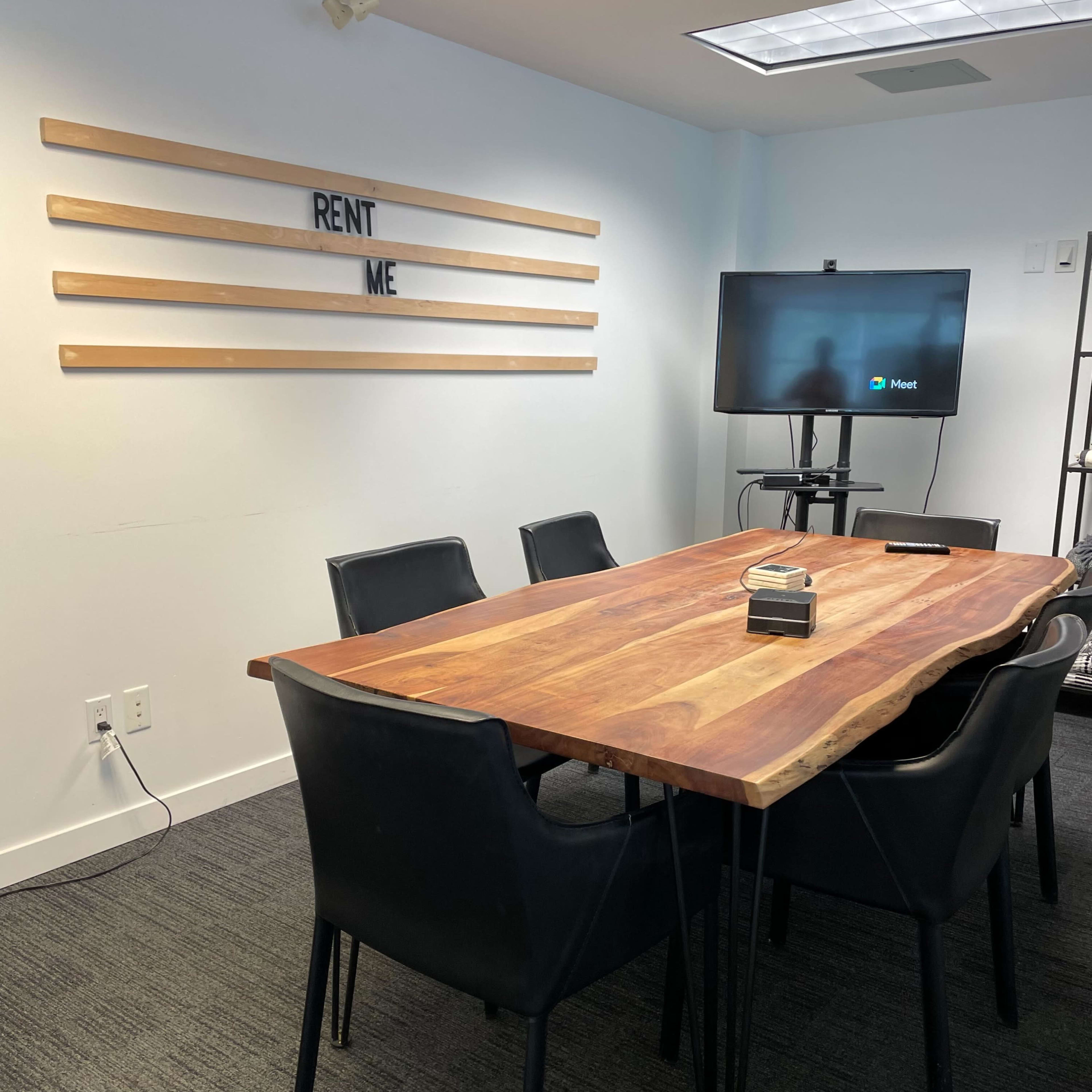 A conference room features a wooden table surrounded by black chairs, with a television and a wall displaying "RENT ME."