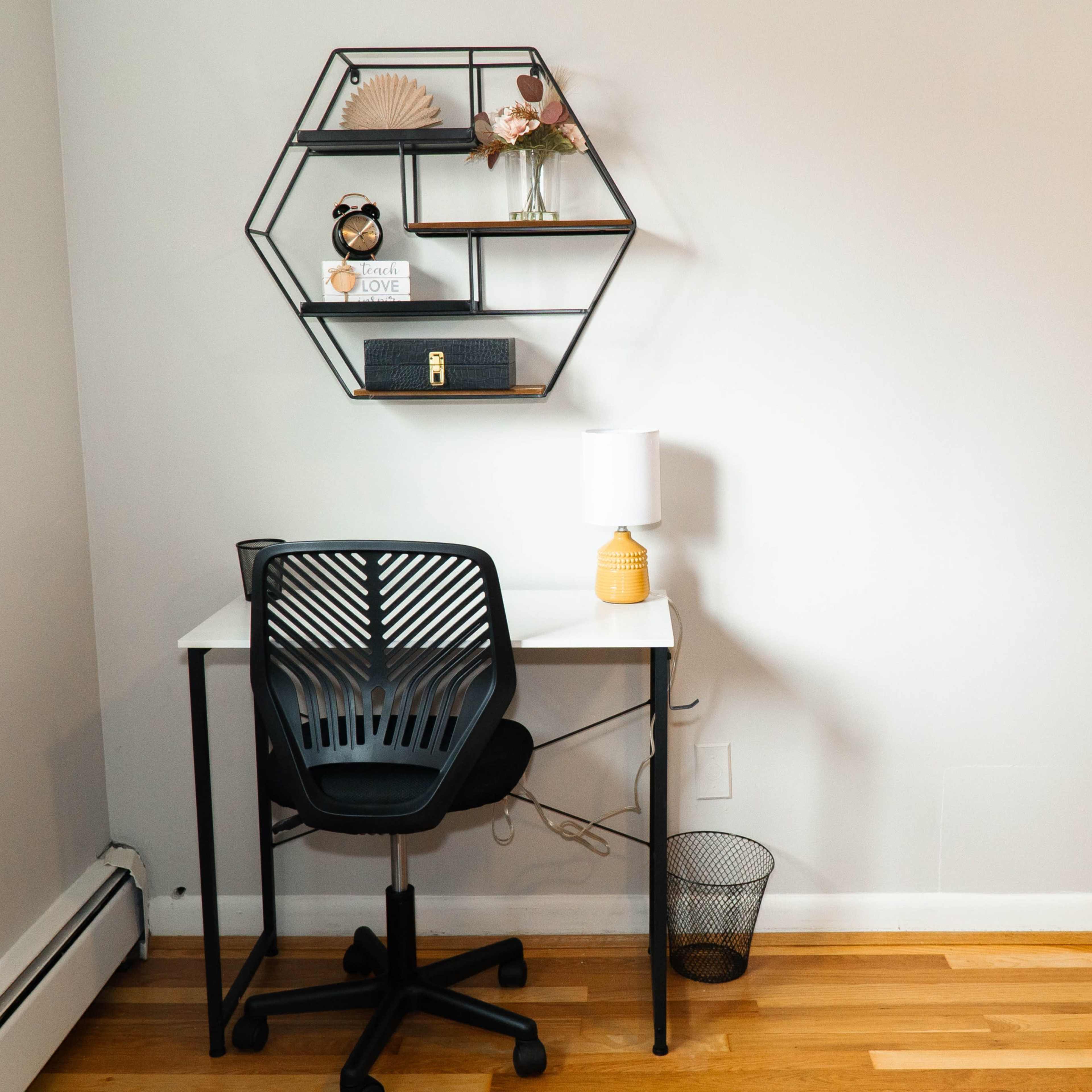 A minimalistic workspace features a desk with a chair, a lamp, and a wall-mounted hexagonal shelf displaying decorative items.