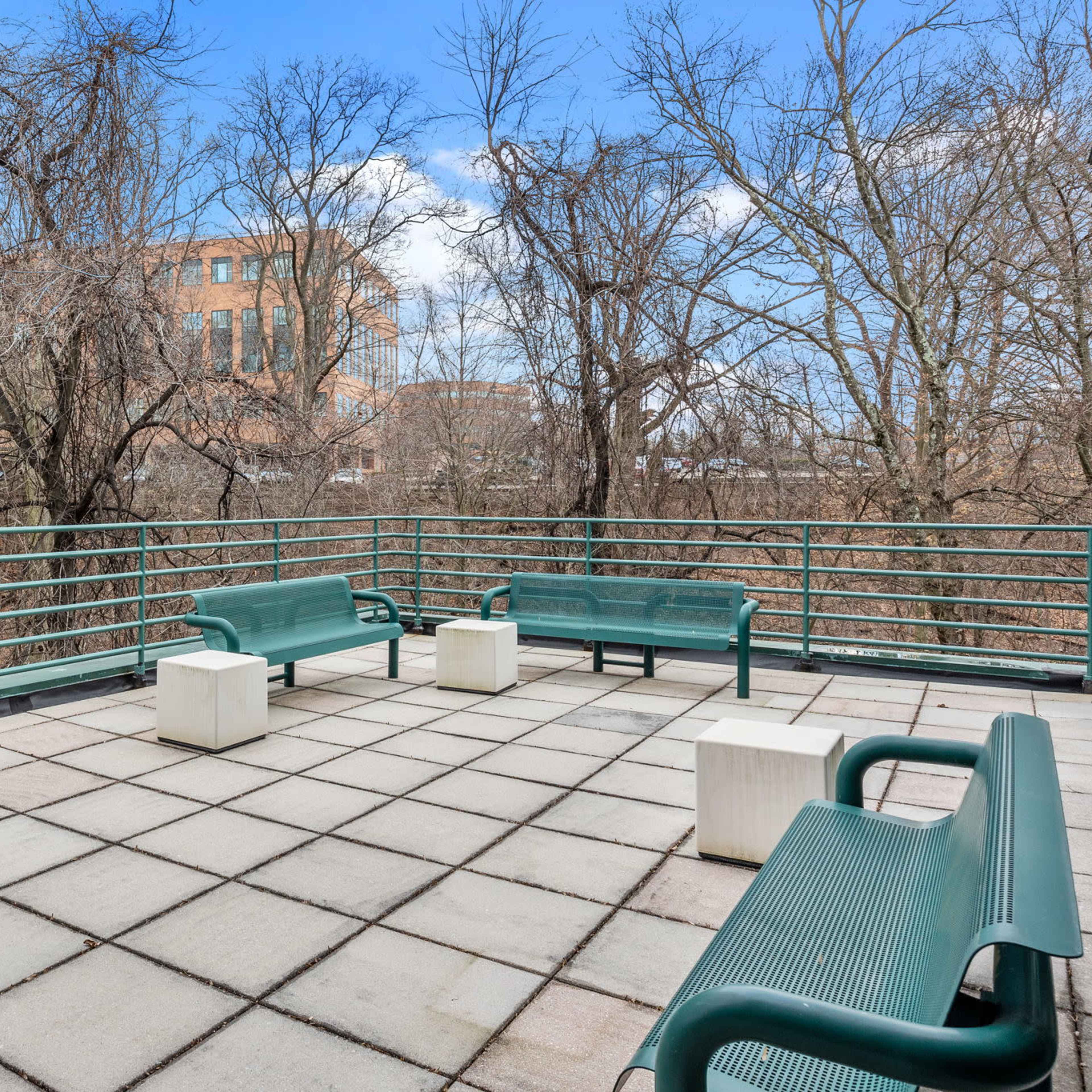 A rooftop terrace features several green benches and concrete tables surrounded by bare trees and a building in the background.