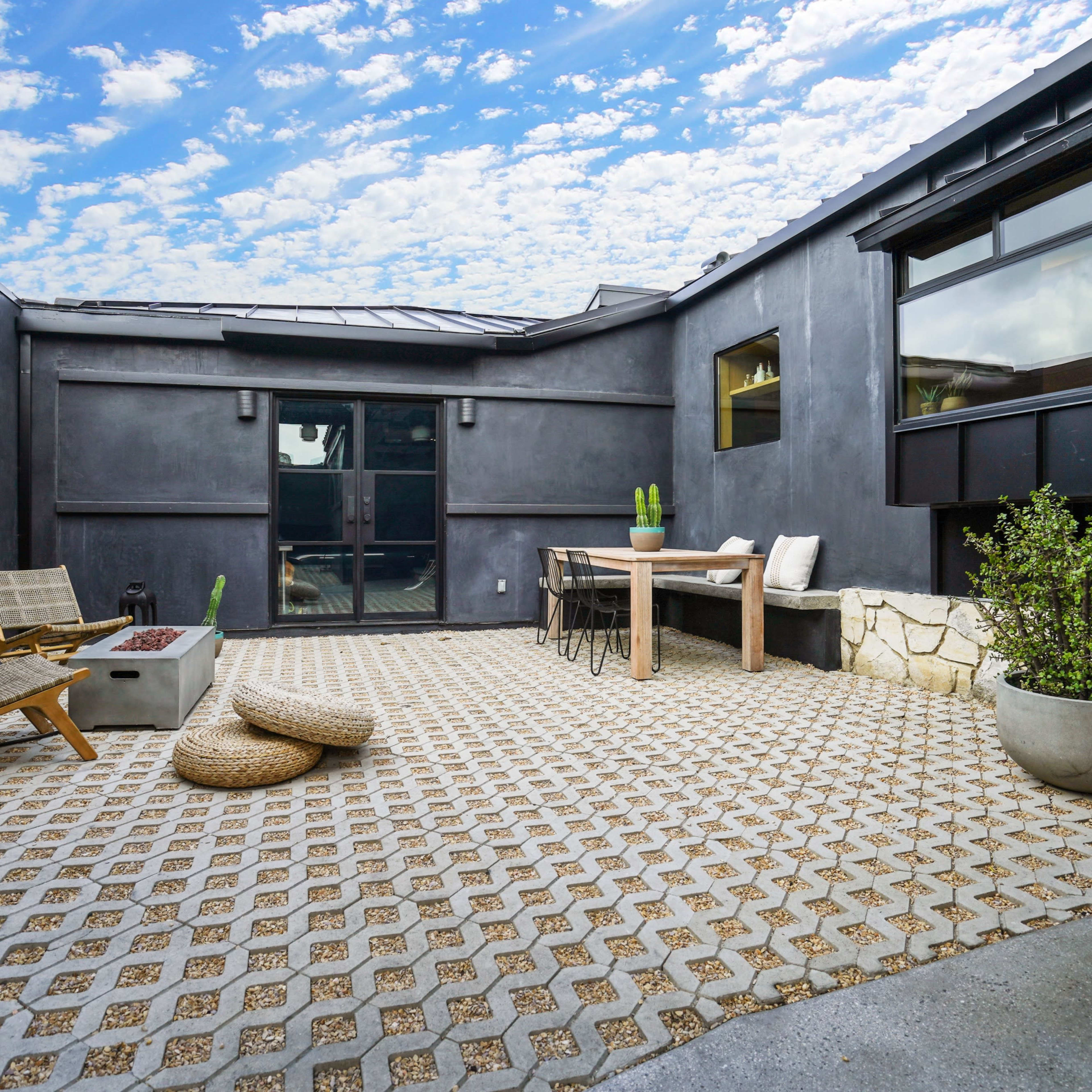 A modern, enclosed courtyard with a patterned stone floor, wooden furniture, and potted plants against black walls and a blue sky.
