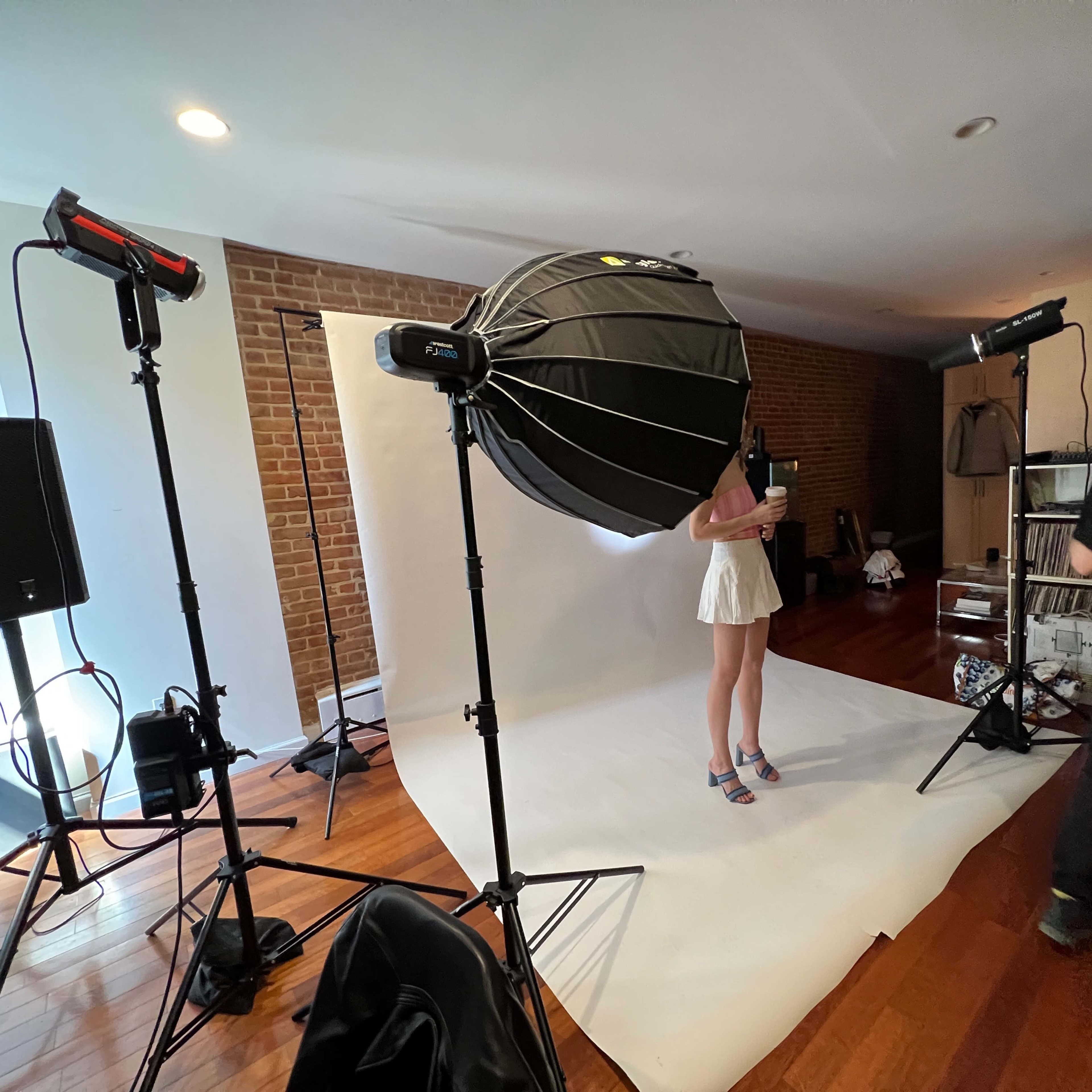 A person in a white dress stands partially obscured behind a large softbox on a photography set with backdrop and lighting equipment.