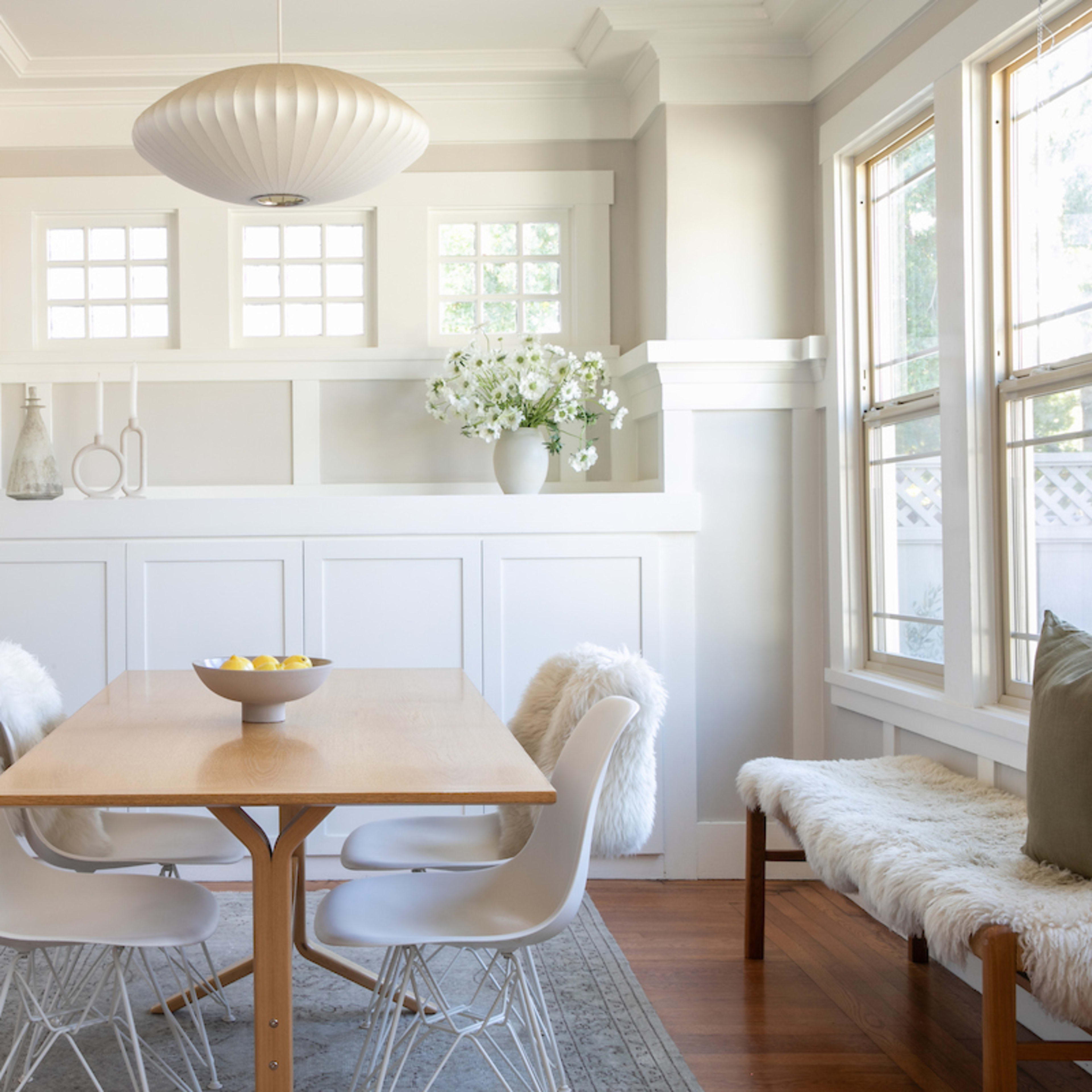 A bright dining area features a wooden table surrounded by four white chairs, with a modern pendant light overhead and a decorative arrangement of flowers and fruit.