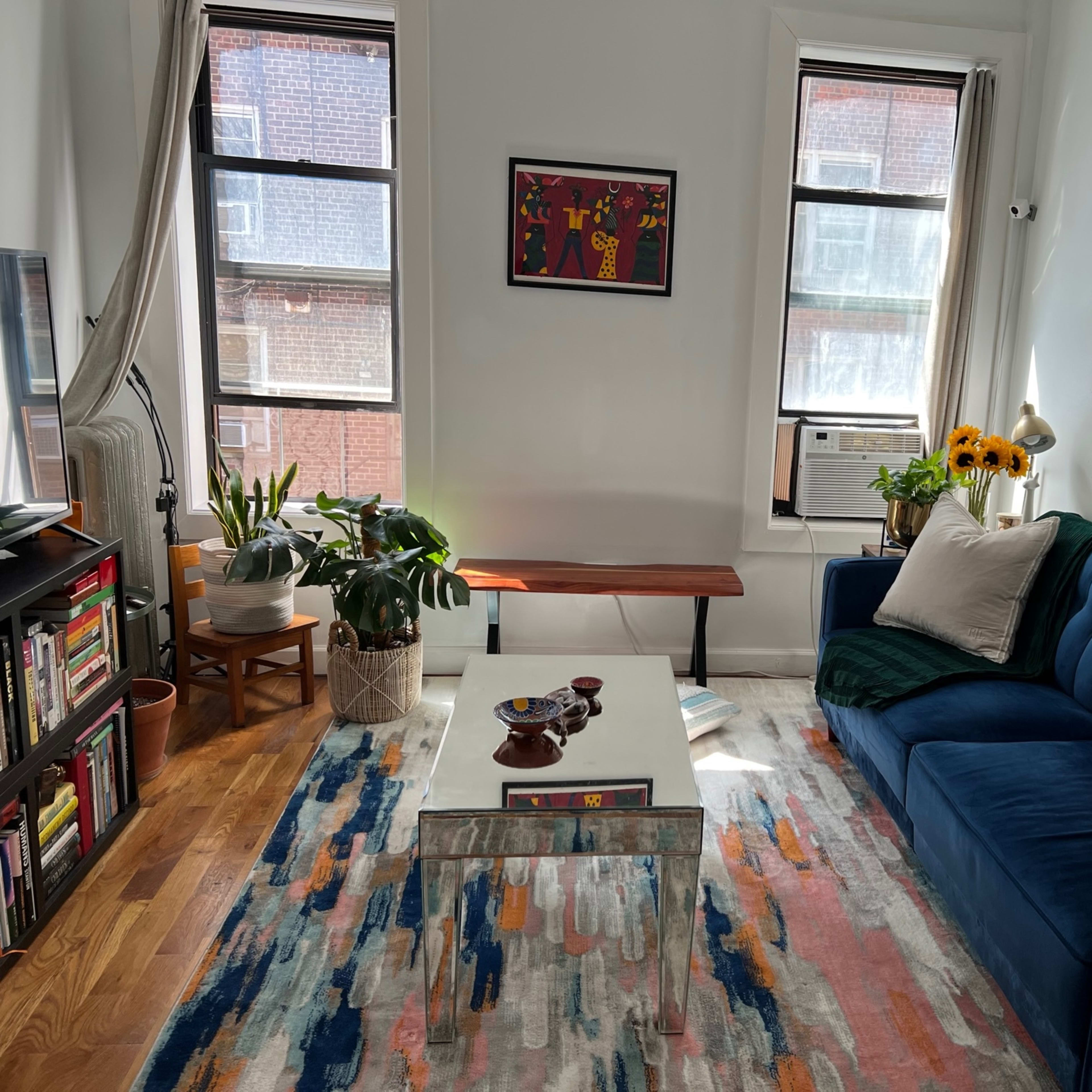 A living room with a blue sofa, a glass coffee table, a colorful rug, potted plants, and framed artwork on the walls.